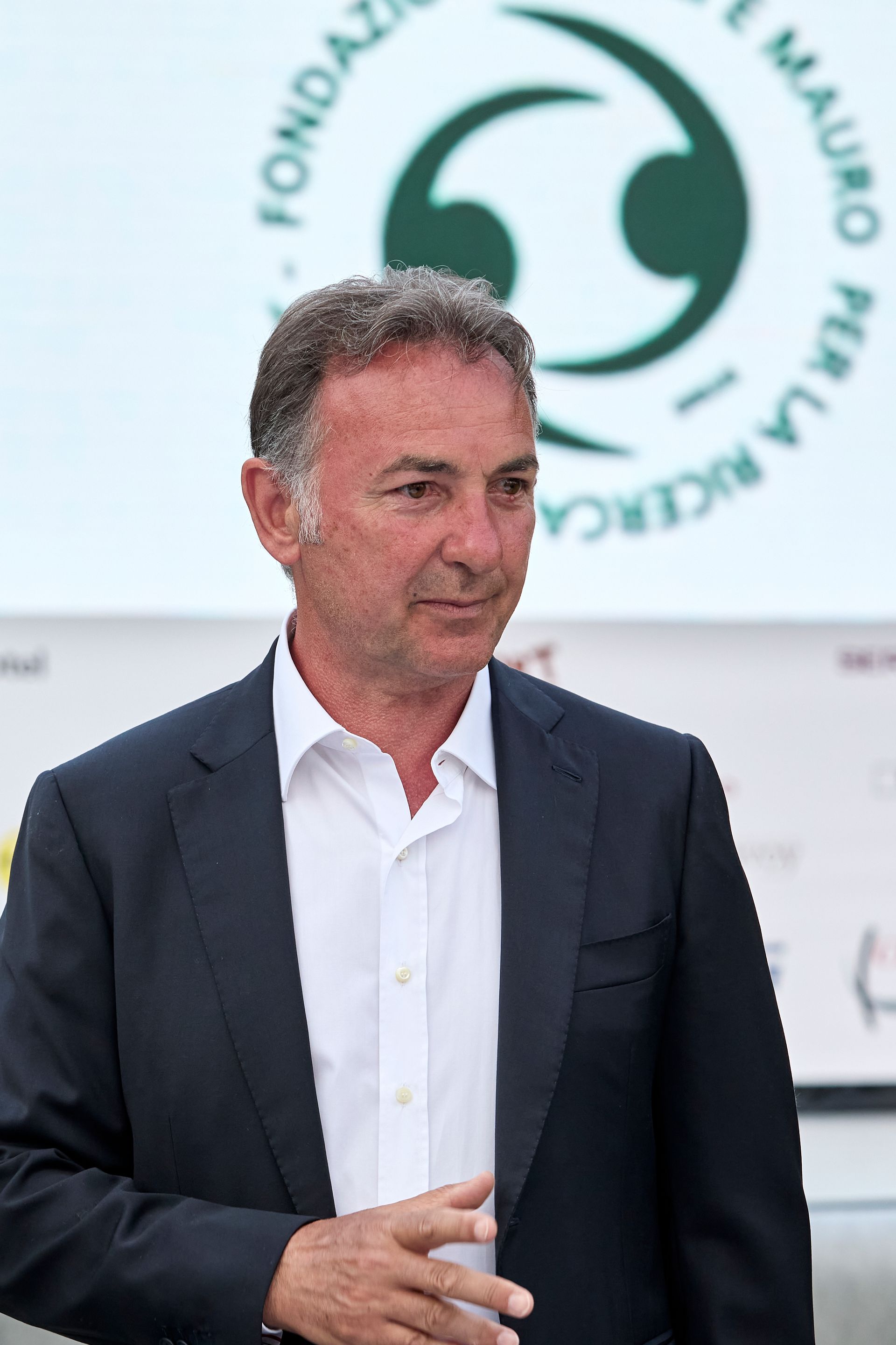 Un uomo in abito scuro e camicia bianca aperta, in piedi all'esterno con un logo visibile sullo sfondo.