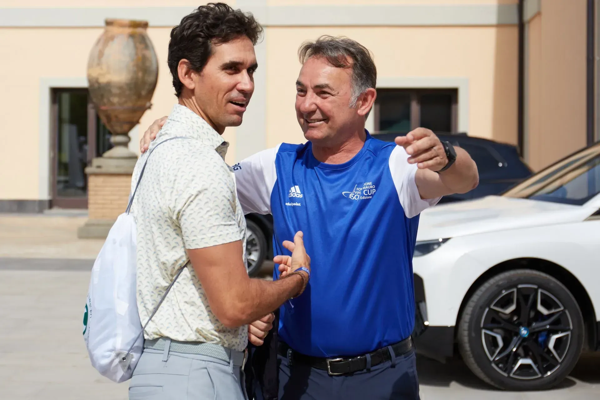 Due uomini alla luce del sole, uno abbraccia l'altro. Un'auto bianca e un edificio beige sullo sfondo.