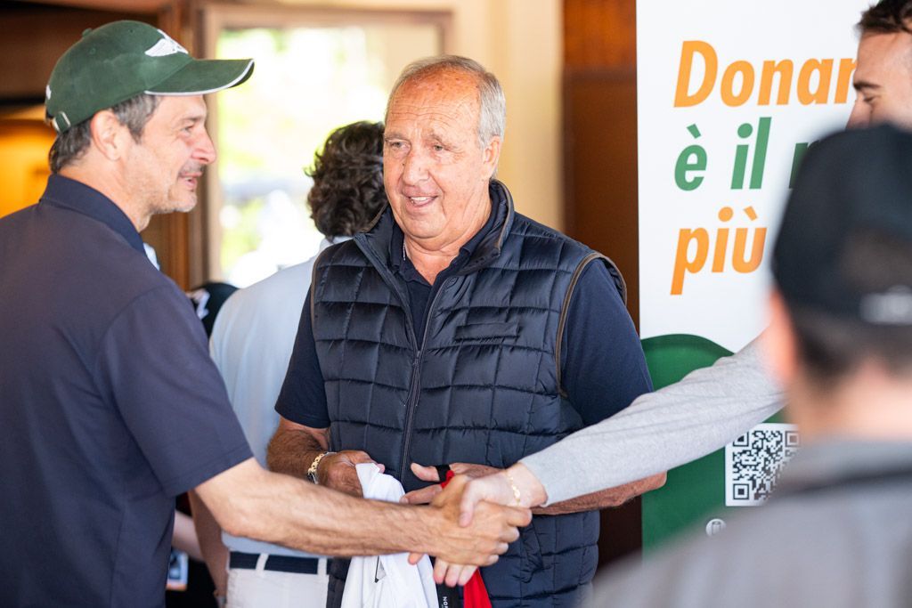 Due uomini a un evento all'aperto. Uno posa in un ritaglio di carta, con il pugno alzato. L'altro è in piedi, sorridente, con indosso un completo da golf.