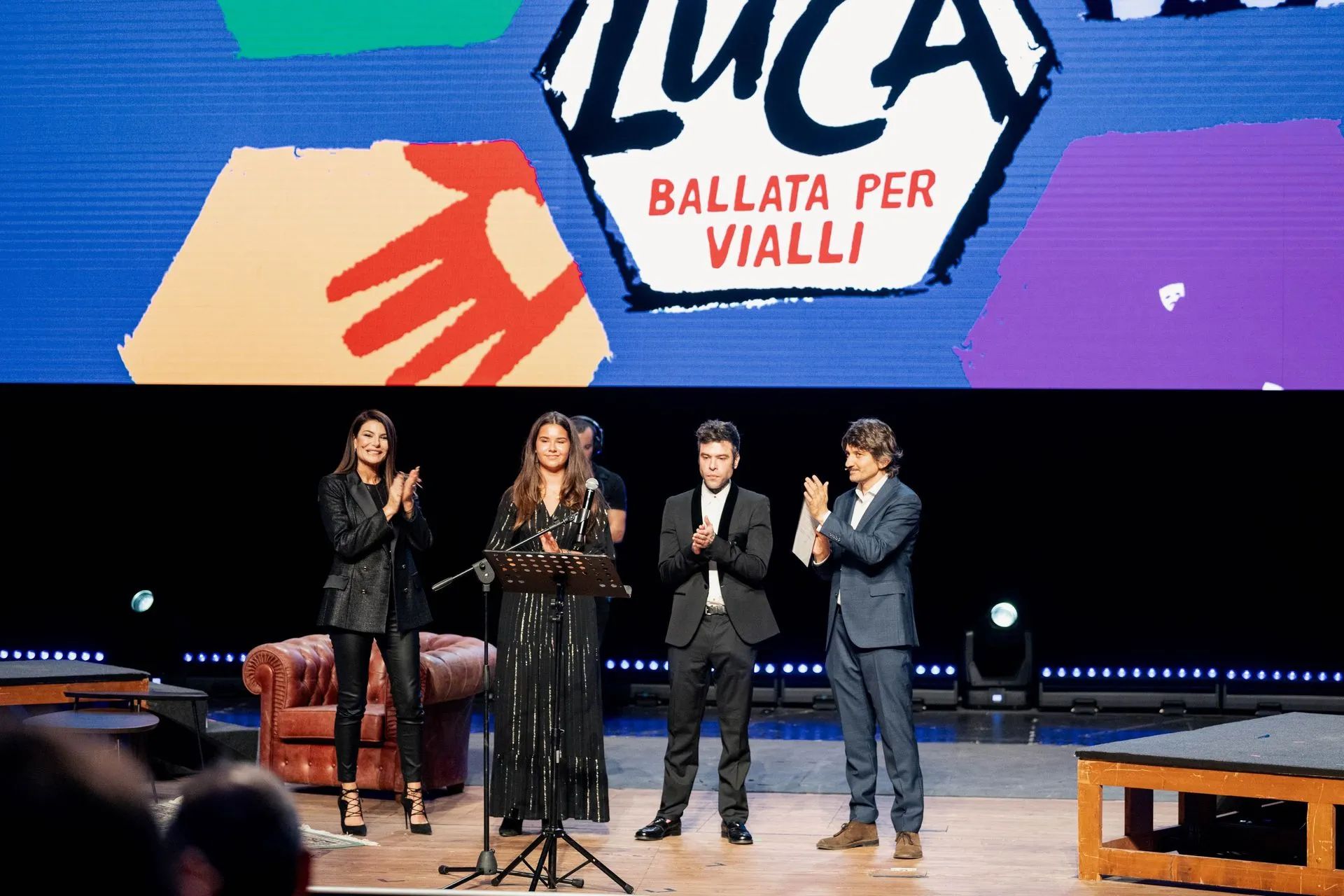 Tre persone sul palco, con microfoni e documenti in mano, davanti allo sfondo con la scritta 