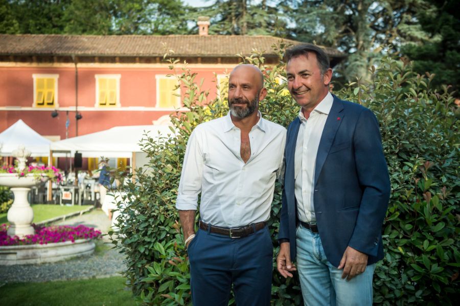 Due uomini posano fuori da un edificio con cortile; uno indossa una camicia bianca, l'altro un blazer blu.
