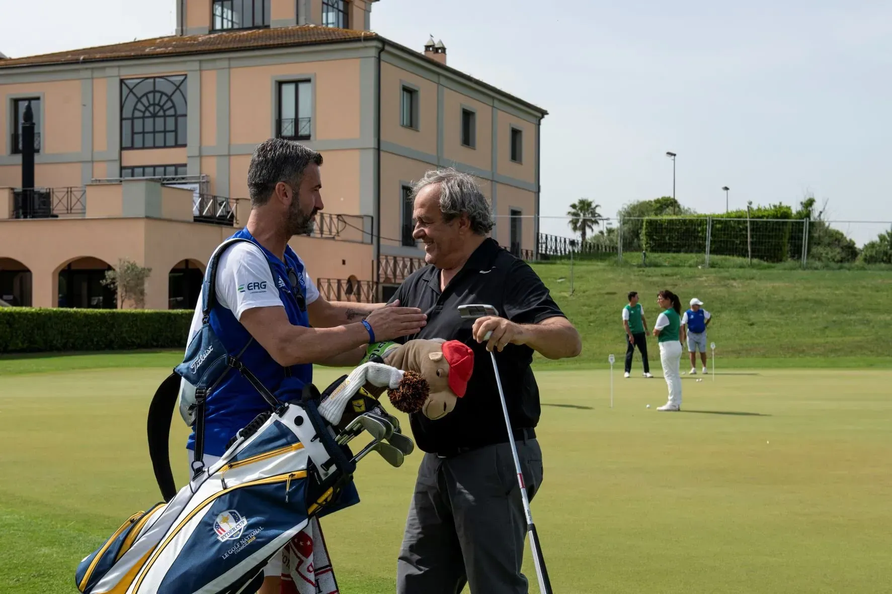 Uomo sul campo da golf con caddy; il caddy gli dà una pacca sul petto. Sacca da golf, erba verde e un edificio sullo sfondo.