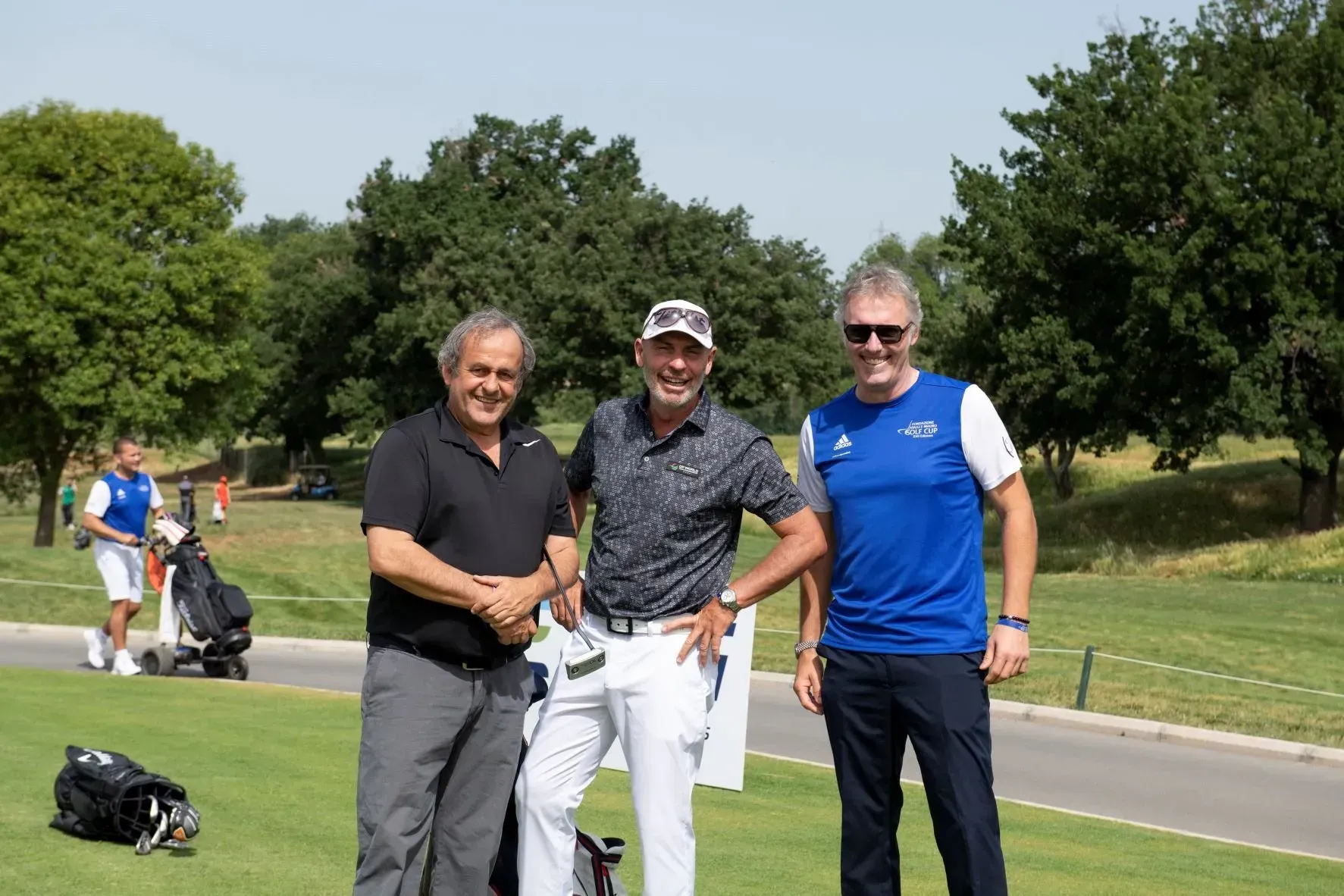 Tre uomini in posa su un campo da golf, due in abiti casual, uno con una maglietta sportiva blu. Erba verde e alberi sullo sfondo.