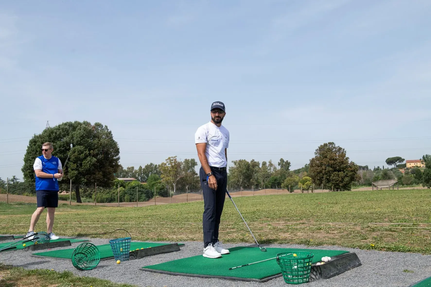 Un uomo si prepara a colpire con una mazza da golf in un campo pratica, mentre un'altra persona è in piedi nelle vicinanze. Scenario soleggiato all'aperto.