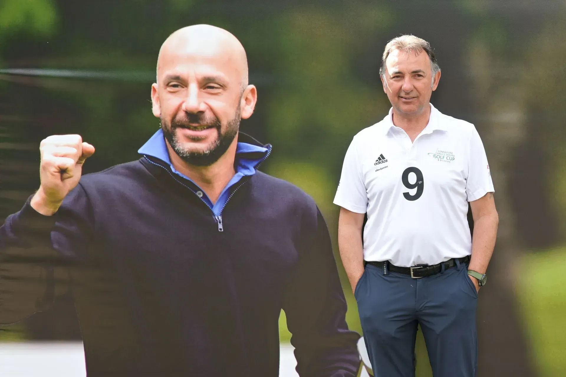 Due uomini a un evento all'aperto. Uno posa in un ritaglio di carta, con il pugno alzato. L'altro è in piedi, sorridente, con indosso un completo da golf.