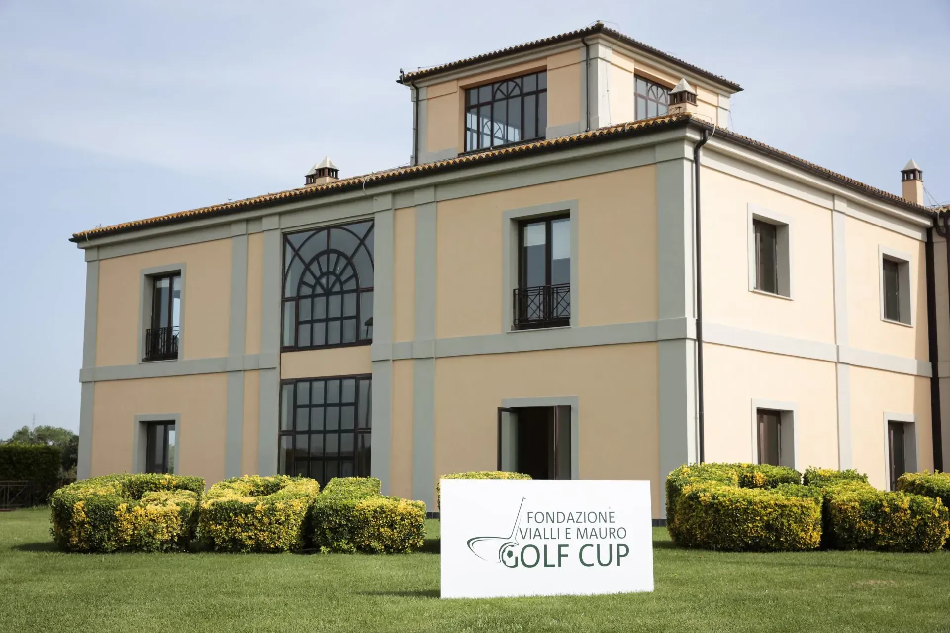 Edificio giallo chiaro con ampie finestre, cartello dell'Italian Open Golf Cup sulla facciata, prato verde.
