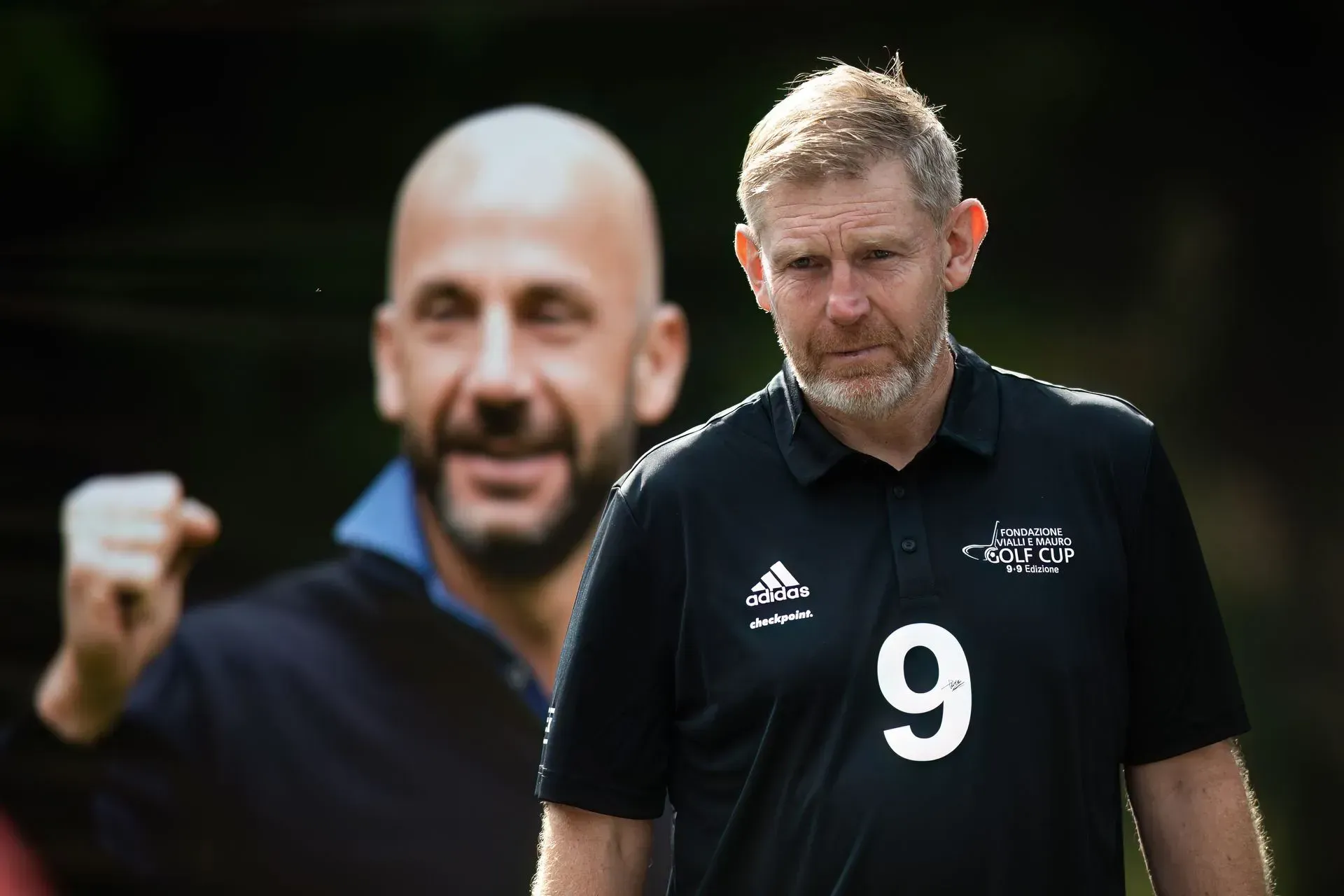 Uomo con camicia nera e numero 9, sullo sfondo sfocato un uomo con la testa rasata.