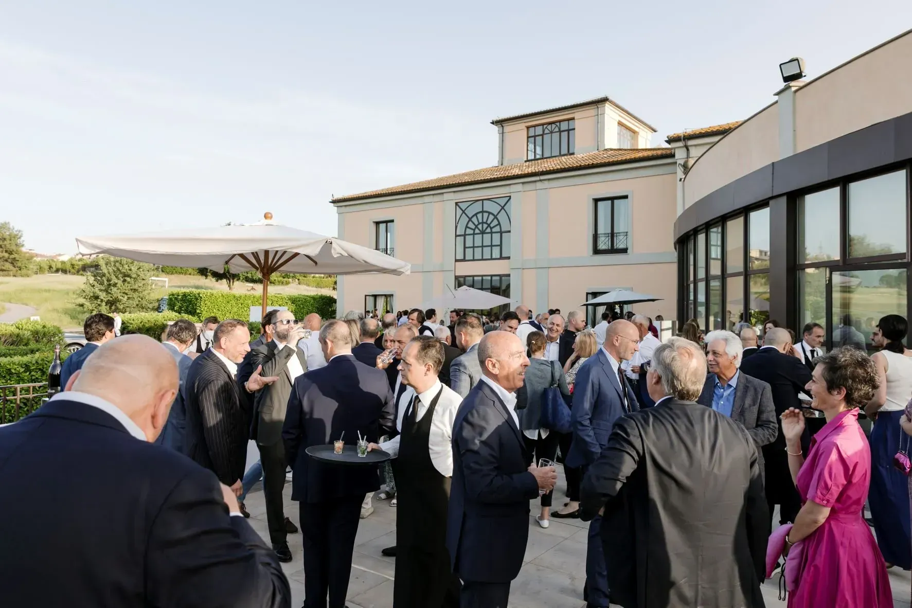 Grande raduno all'aperto: persone in giacca e cravatta socializzano, sorseggiando un drink, vicino a un edificio con terrazza.