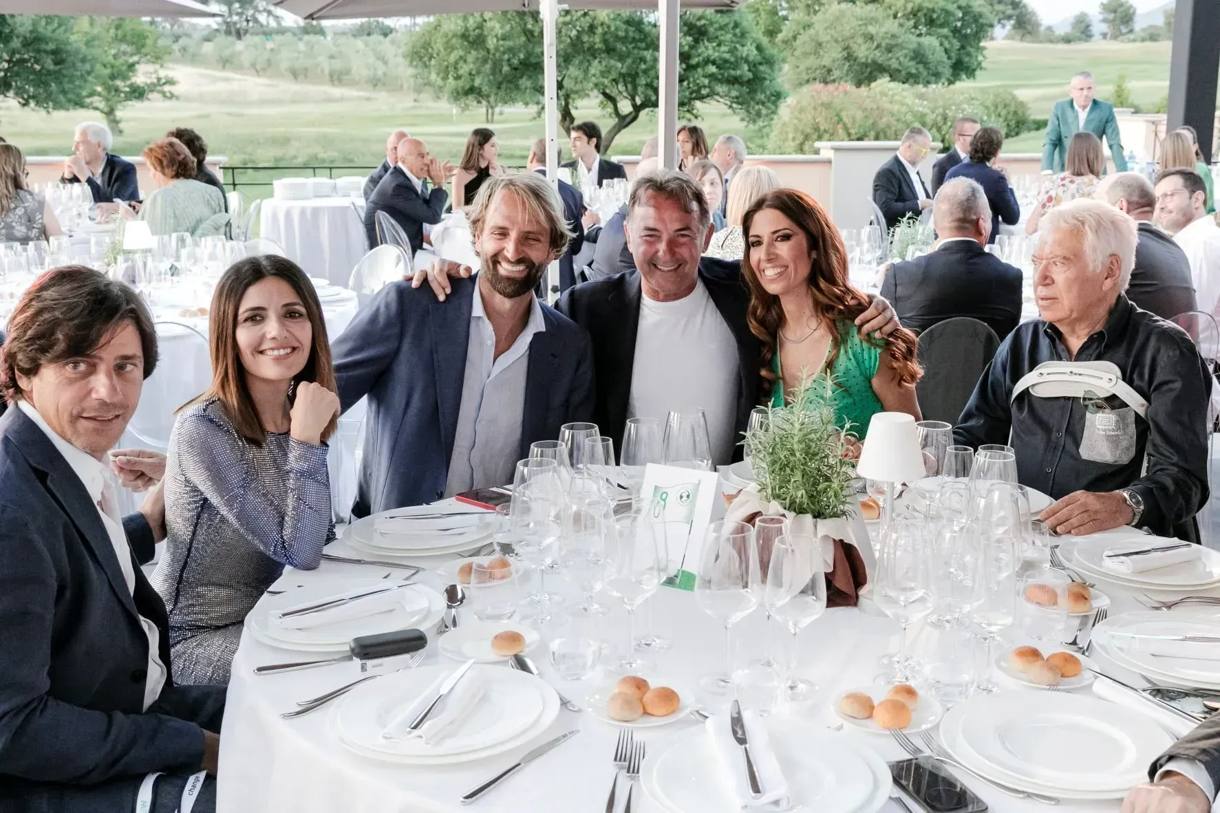 Un gruppo di persone sorridenti attorno a un tavolo rotondo apparecchiato per un pasto, all'aperto con un campo da golf sullo sfondo.