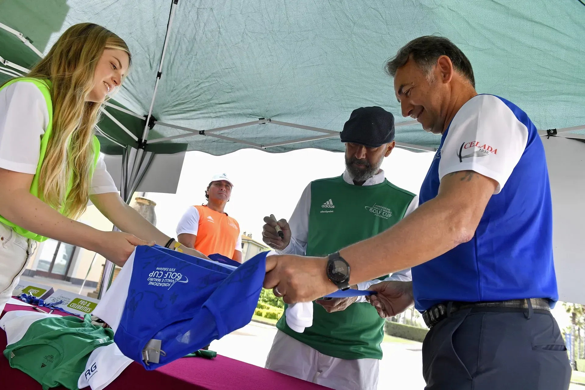 Una donna porge un berretto blu a un uomo, entrambi sotto una tenda. Altri sono lì vicino durante un evento di golf.