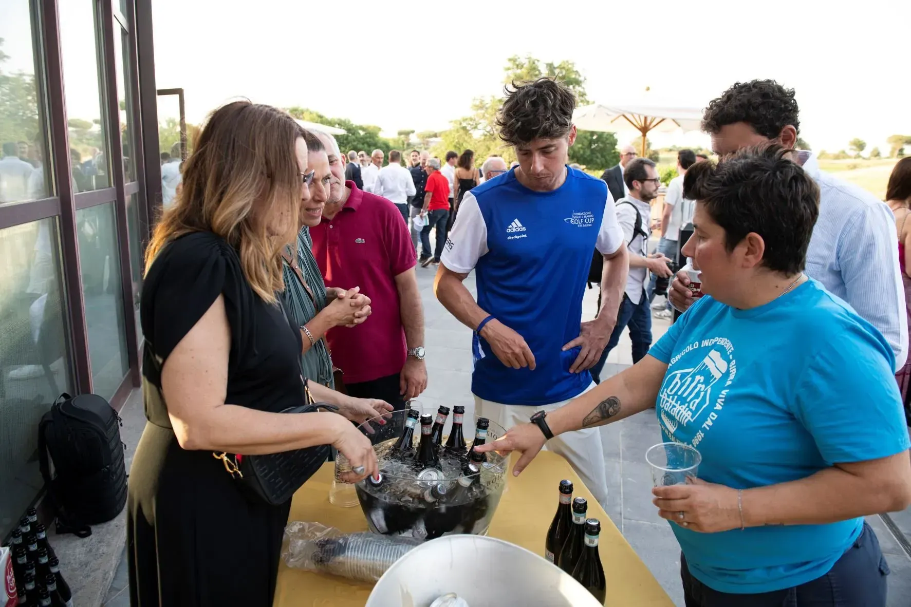 Persone riunite a un evento all'aperto, intente a scegliere le bevande. Una donna con una camicia blu indica le bottiglie su un tavolo.