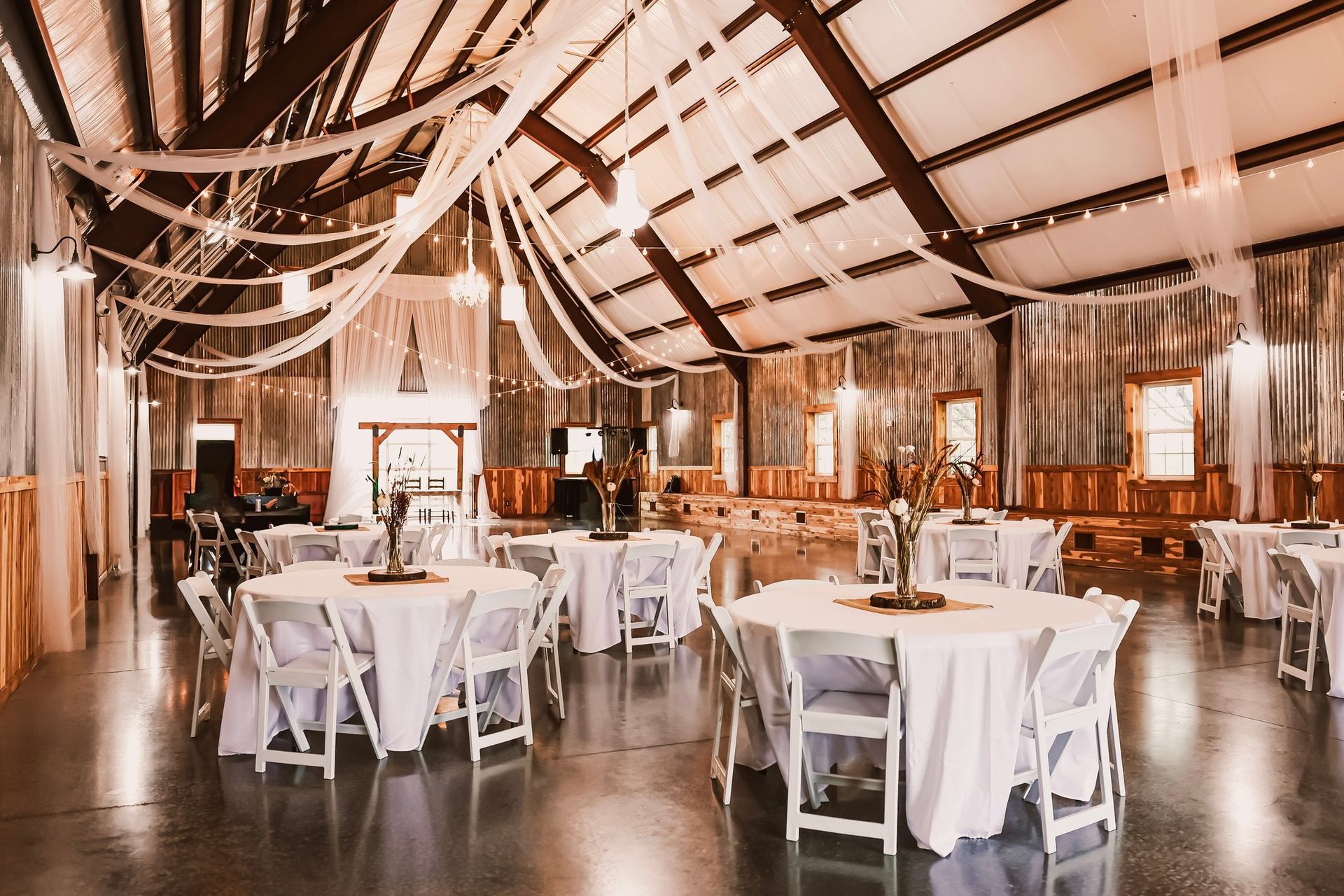 Reception hall, decorated with white fabric, tables, and chairs. Wooden interior, neutral lighting.