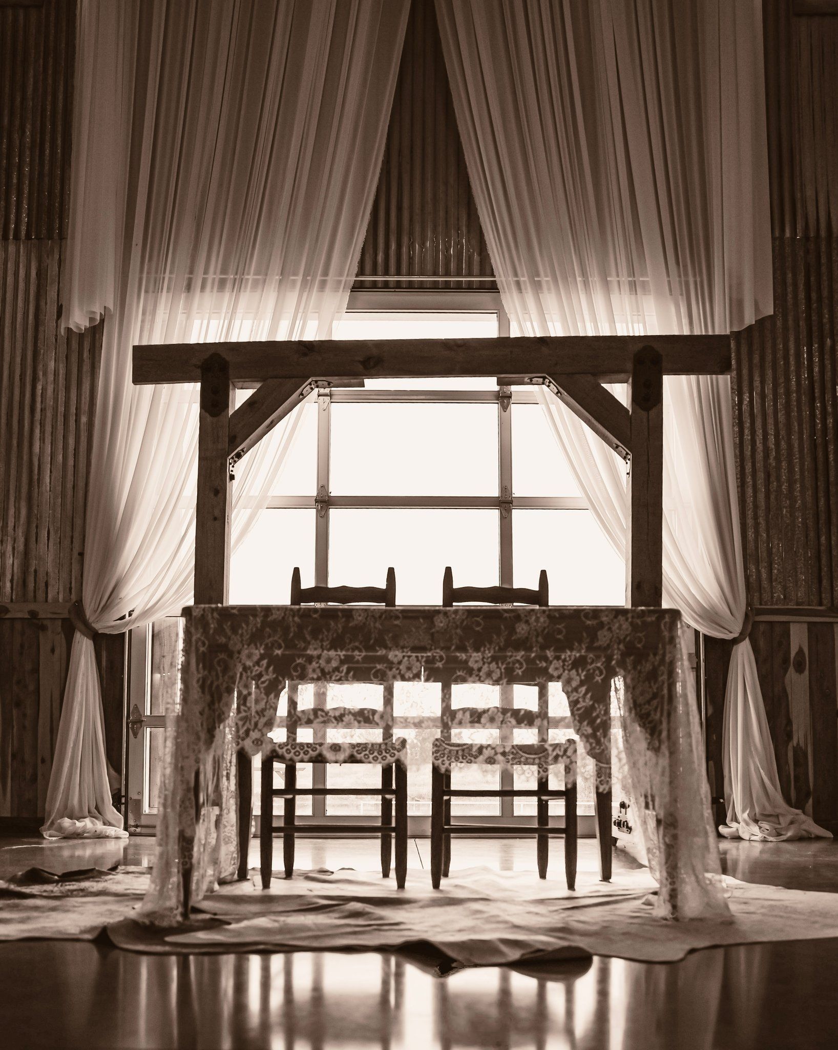 Wedding altar in sepia tone, two chairs facing a window, draped with fabric and under a wooden arch.