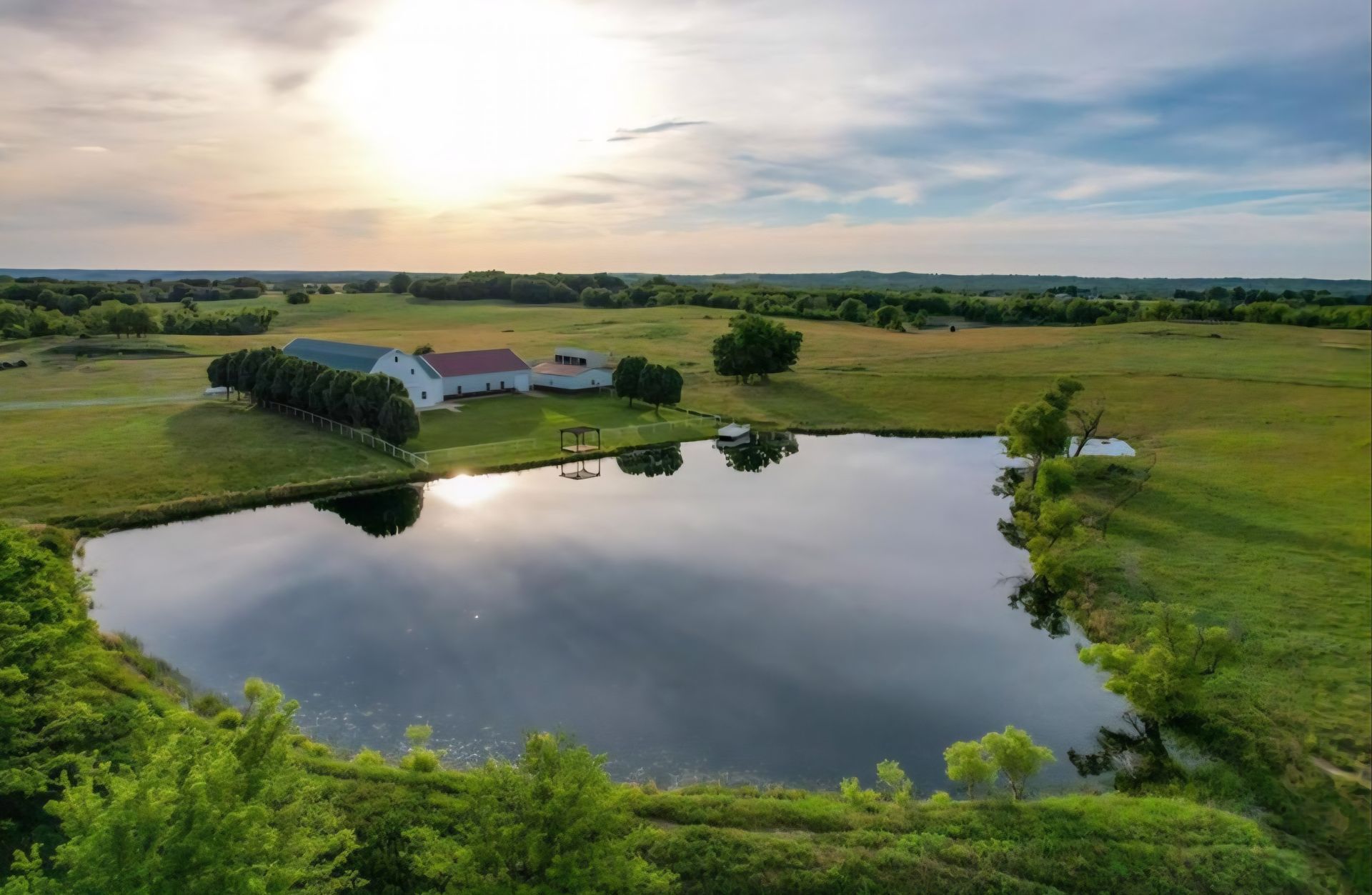 Tranquil farm scene with a pond reflecting the sky, near a white building and green fields at sunset.