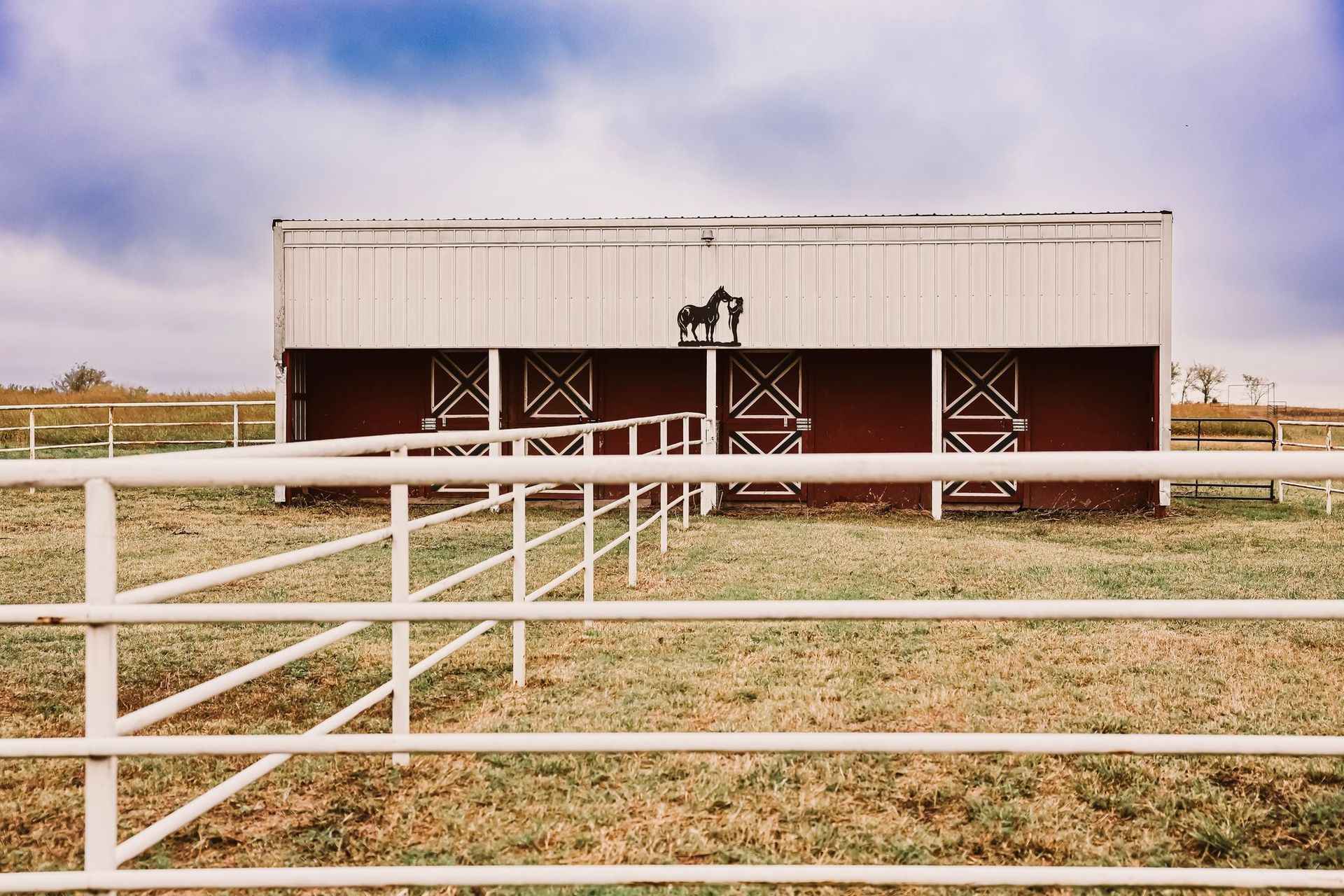 White metal barn with horse statue on top; red doors, white fence, and grassy field.