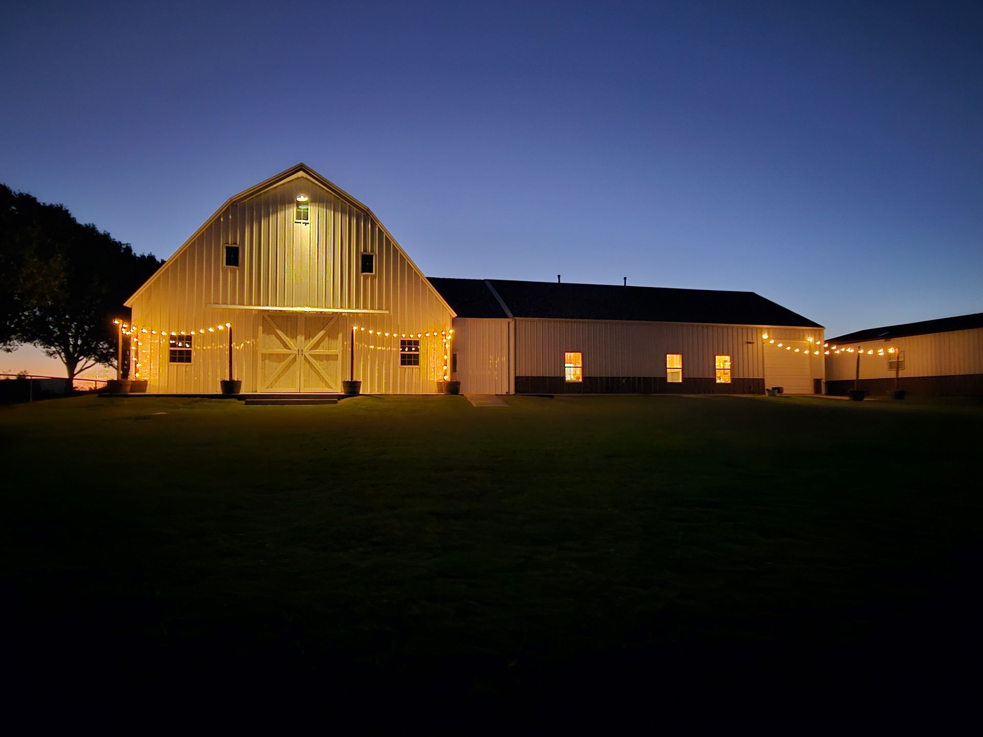 White barn with warm lights against a dusky blue sky. String lights and open doors.