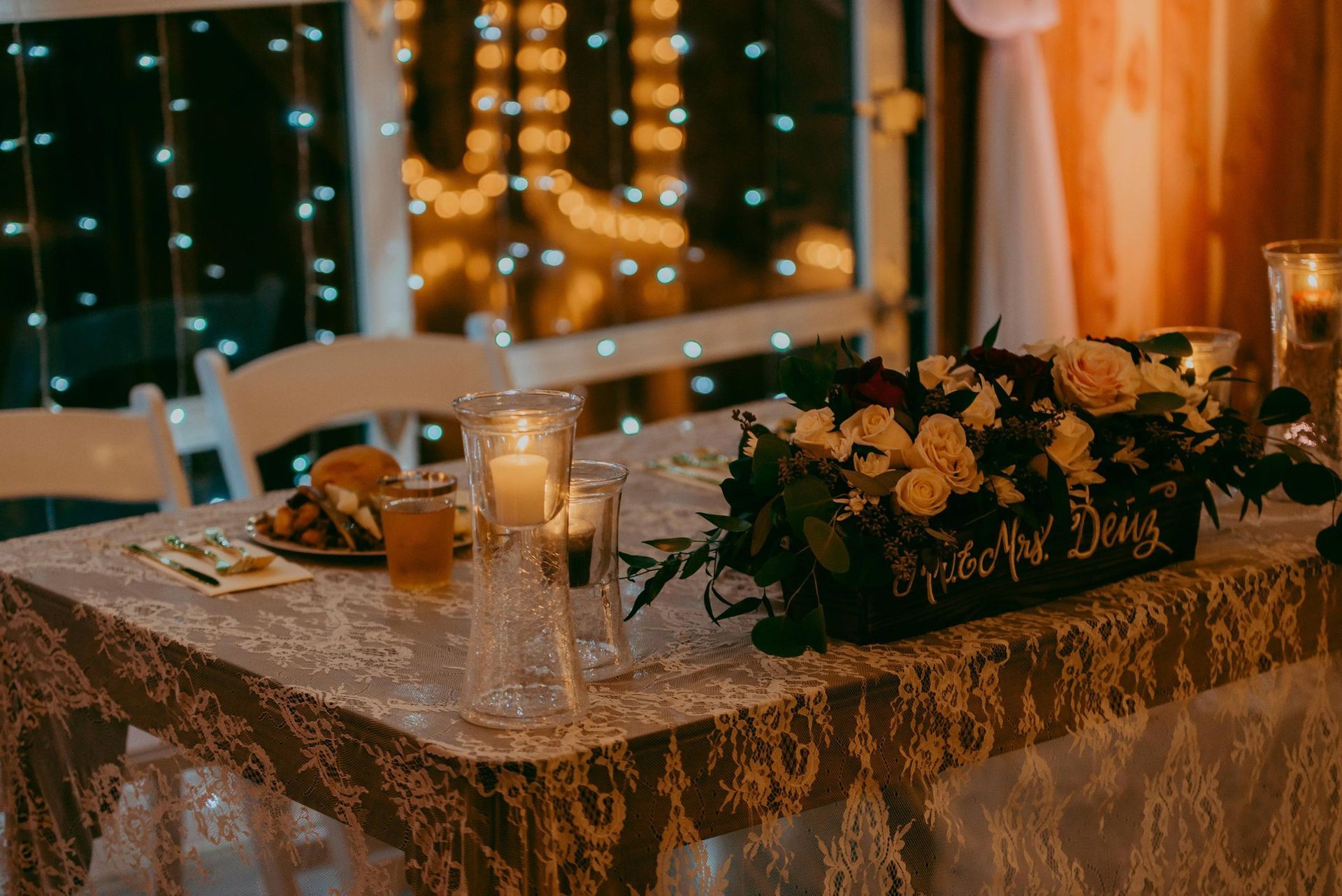Wedding reception table with floral centerpiece, candles, and string lights in the background.