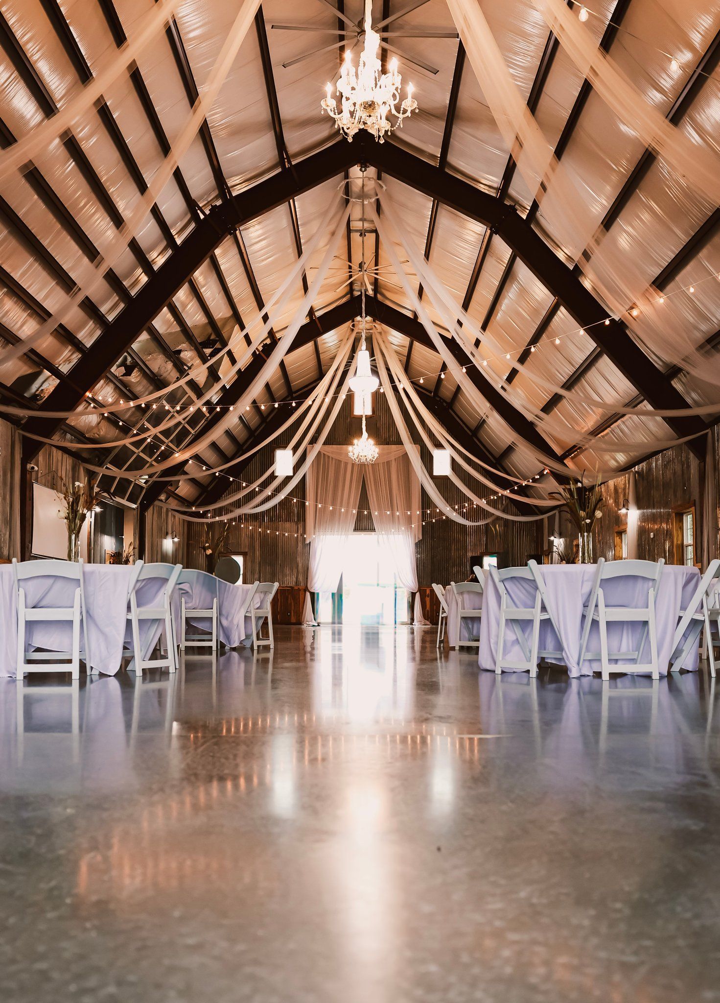 Wedding reception in a barn, decorated with white linens, string lights, and draping fabric.