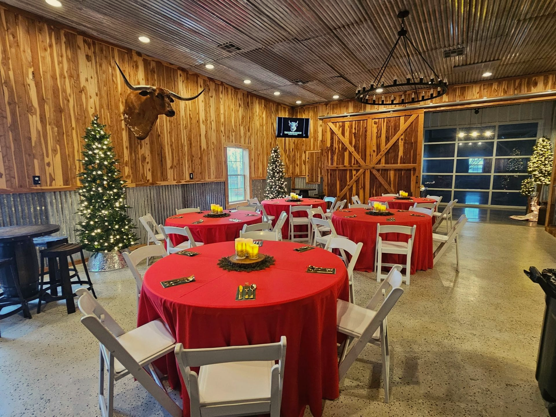 Event space with red-covered round tables, white chairs, Christmas trees, and a longhorn head mount.