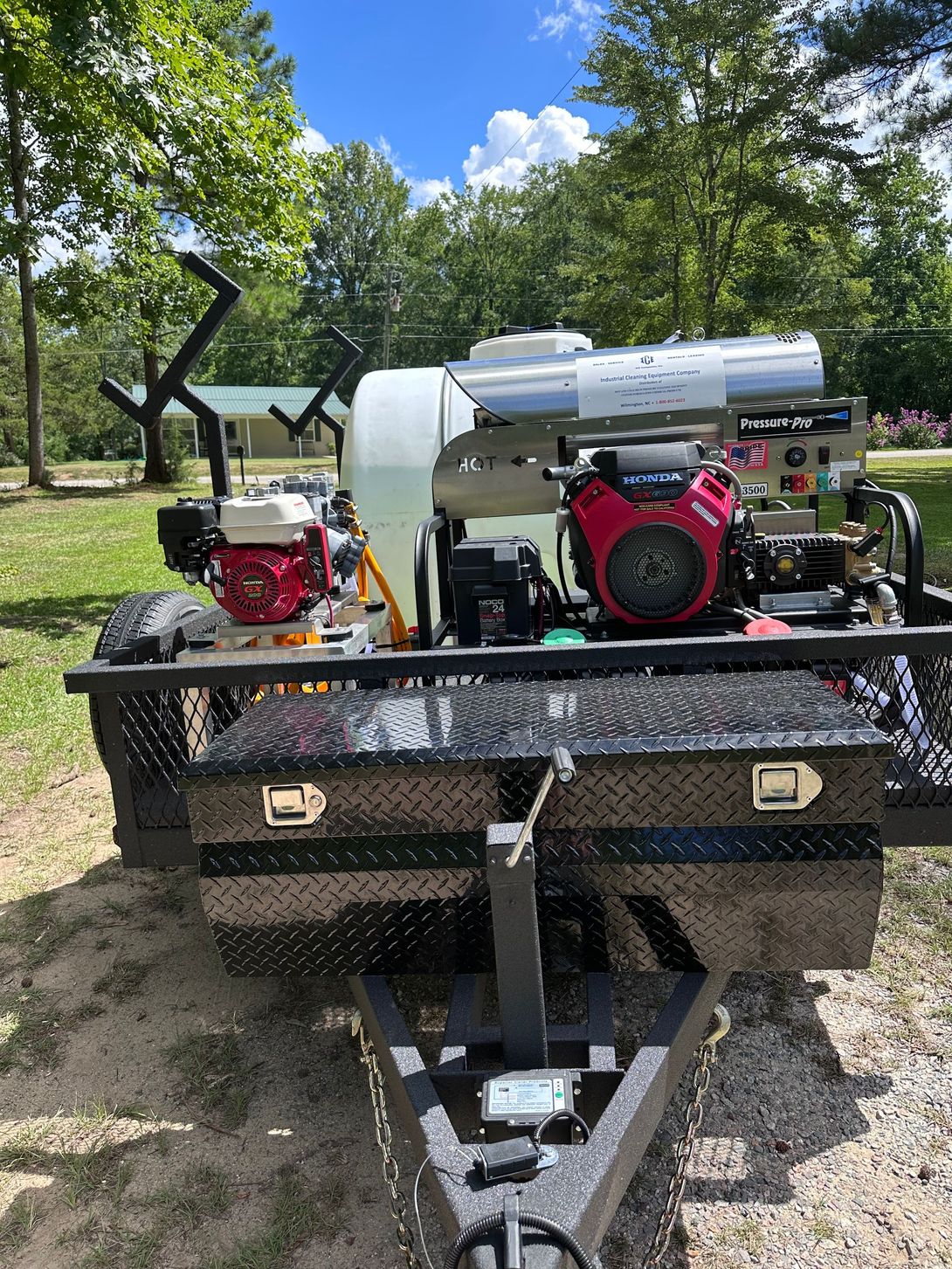 Pressure washing equipment on a trailer. Black and red engines, white water tank, diamond plate toolbox, outdoors.