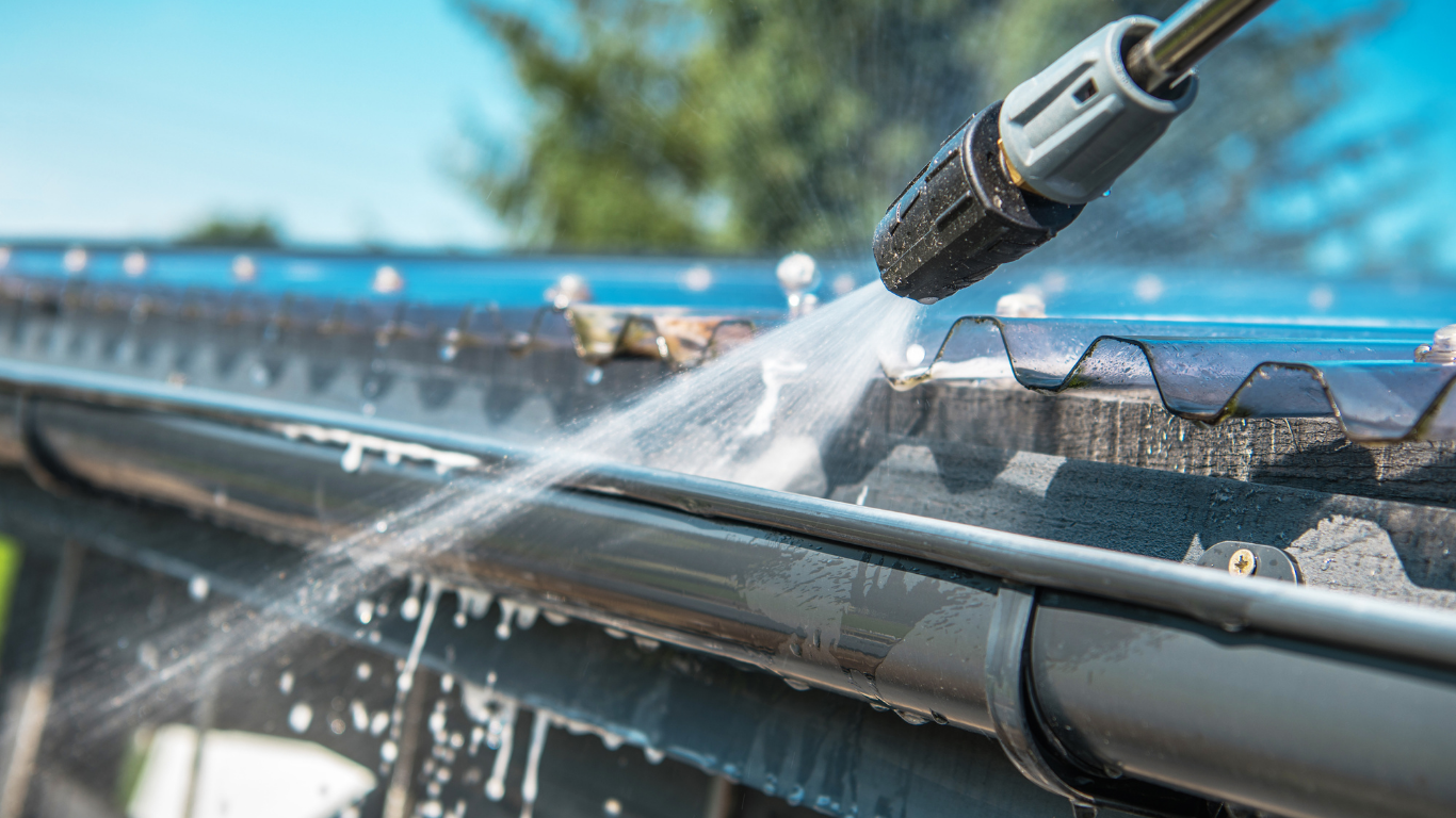 A person using a pressure washer to clean a black gutter on a roof.