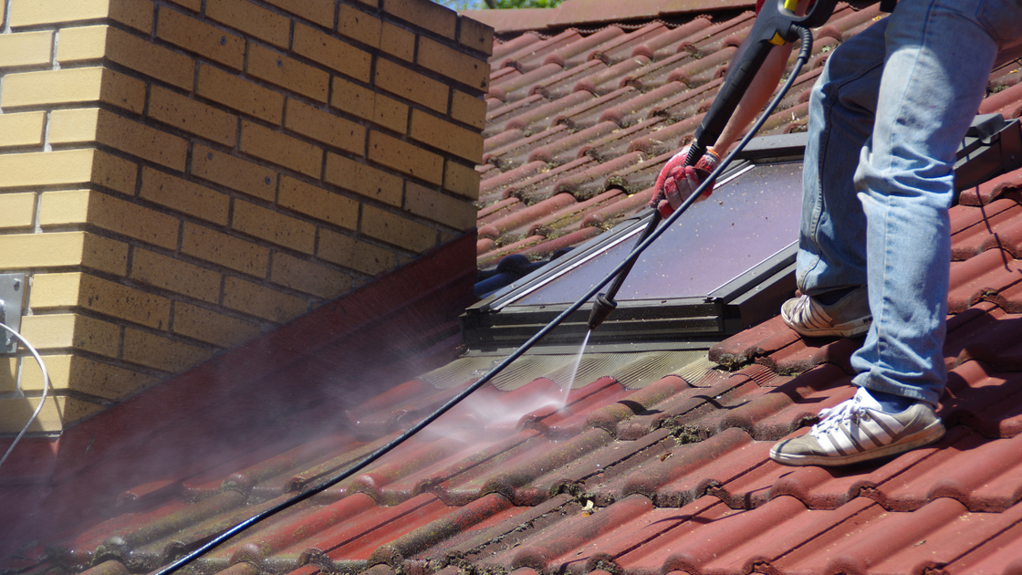 Person power washing a red tile roof near a brick chimney and a skylight.