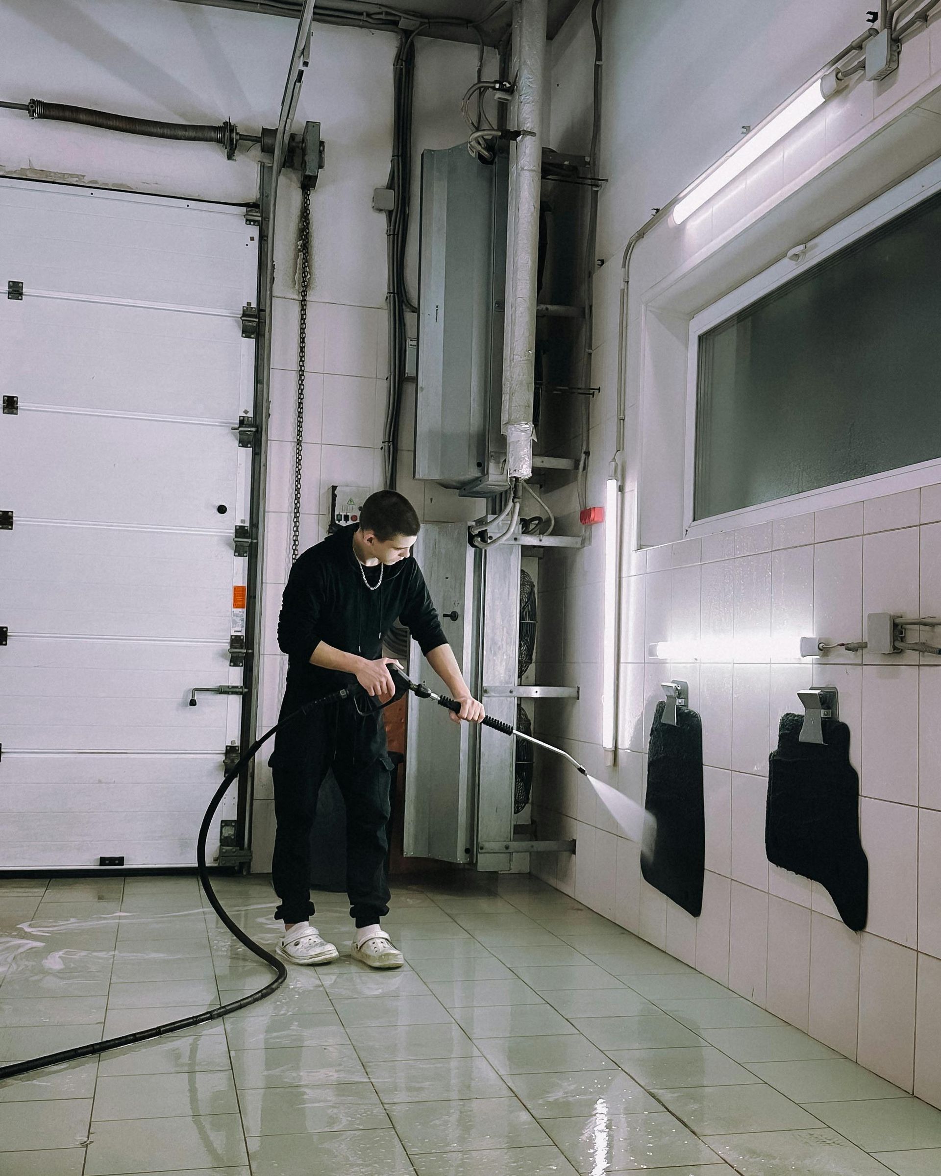 Man cleaning a tiled wall with a pressure washer in a car wash bay.