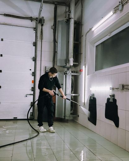 Man washing a tiled wall with a pressure washer in a garage-like setting.
