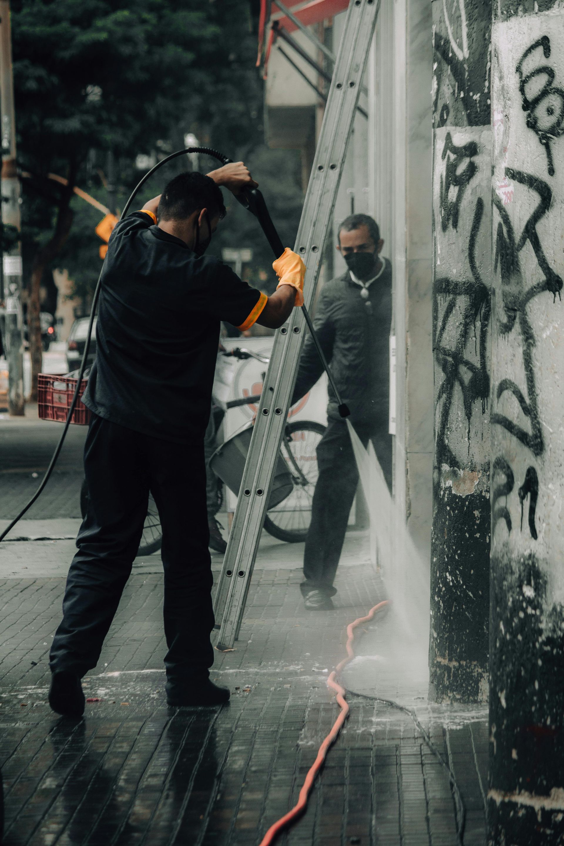 Two people power washing graffiti off a building's exterior wall on a city street.