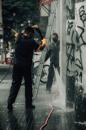 Two people power washing graffiti off a building's exterior wall on a city street.