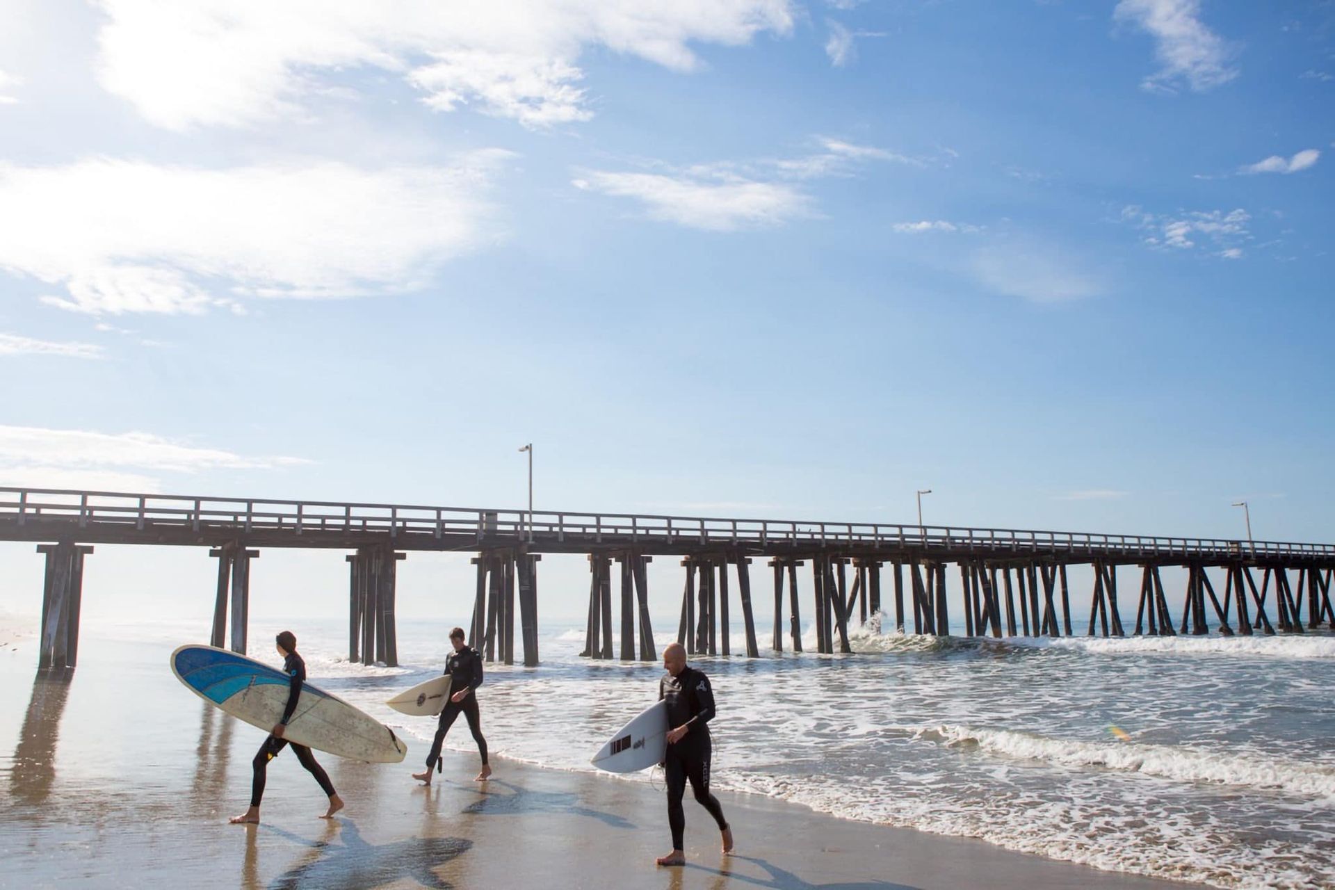 Three surfers are walking on the beach near a pier.