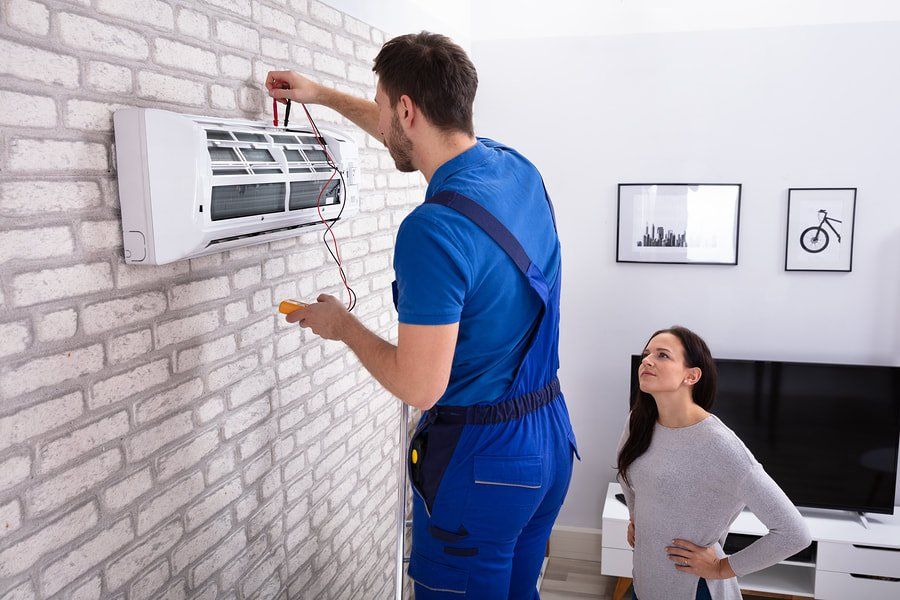 A man is fixing an air conditioner in a living room while a woman looks on.
