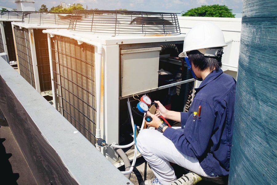 A man is sitting on the roof of a building working on an air conditioner.