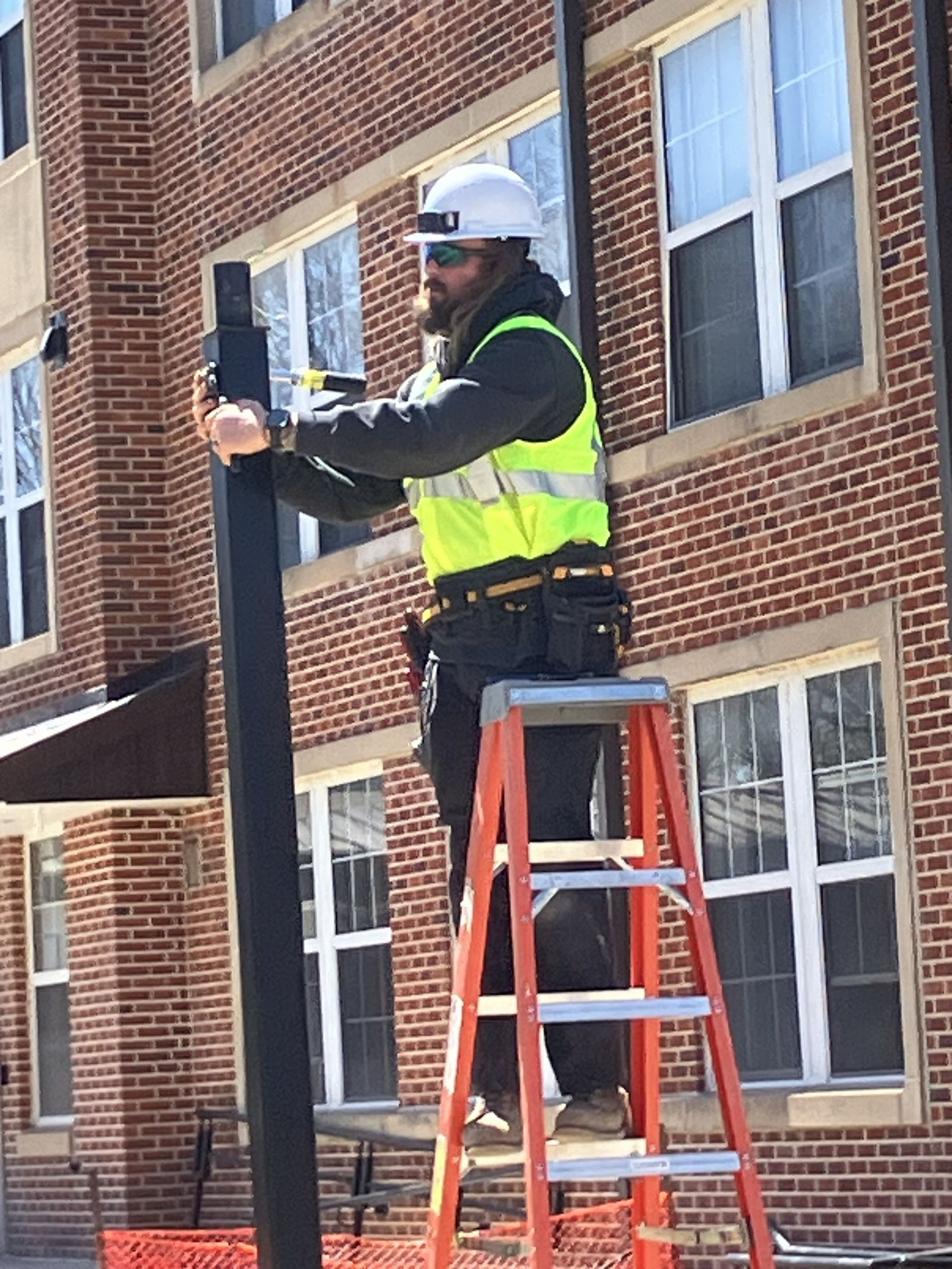 Worker in hard hat and safety vest standing on a ladder beside a brick building, adjusting a pole-mounted device.