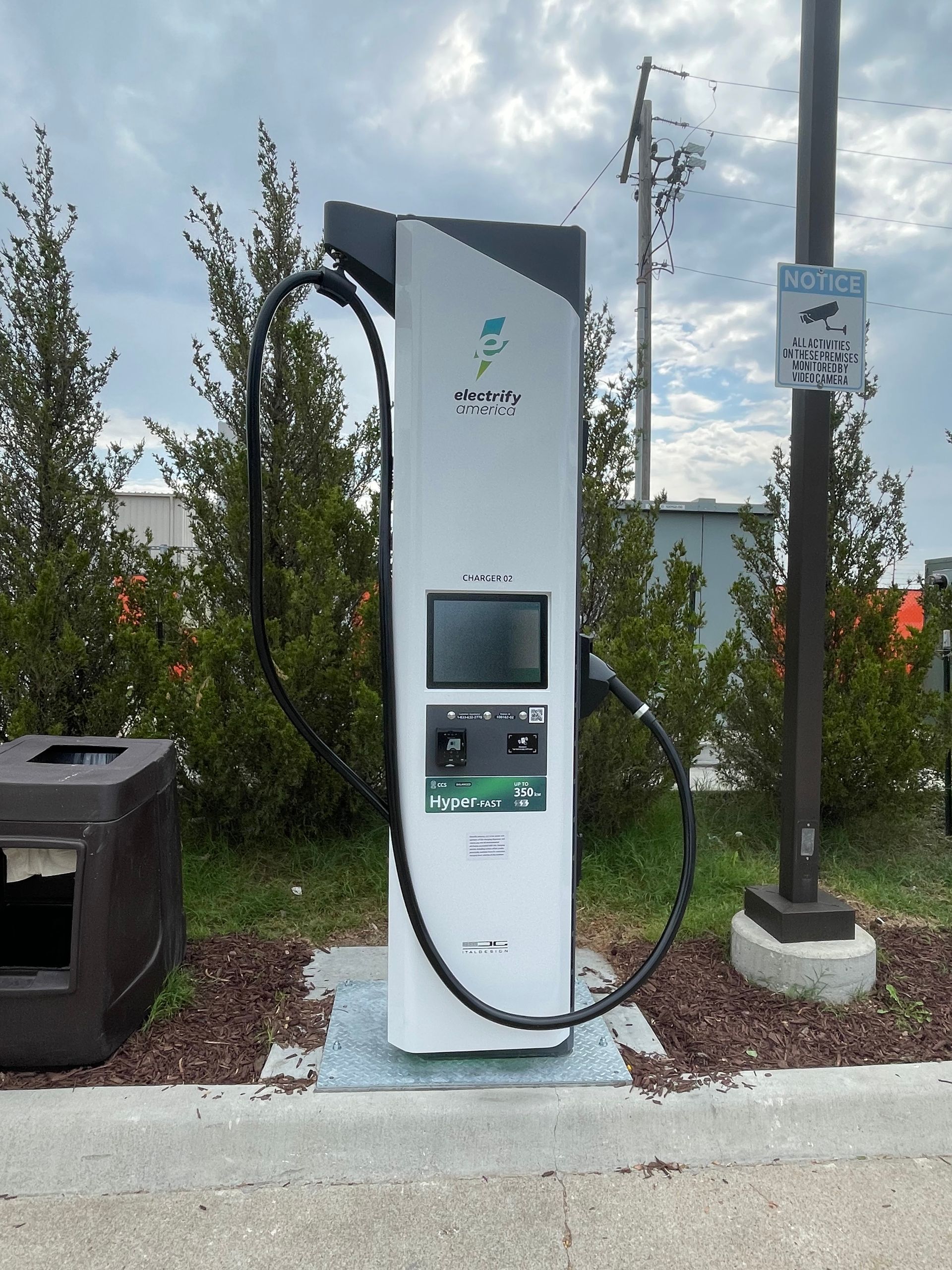 Electric vehicle charging station on a sidewalk beside a parking area, with a white charger and small screen.