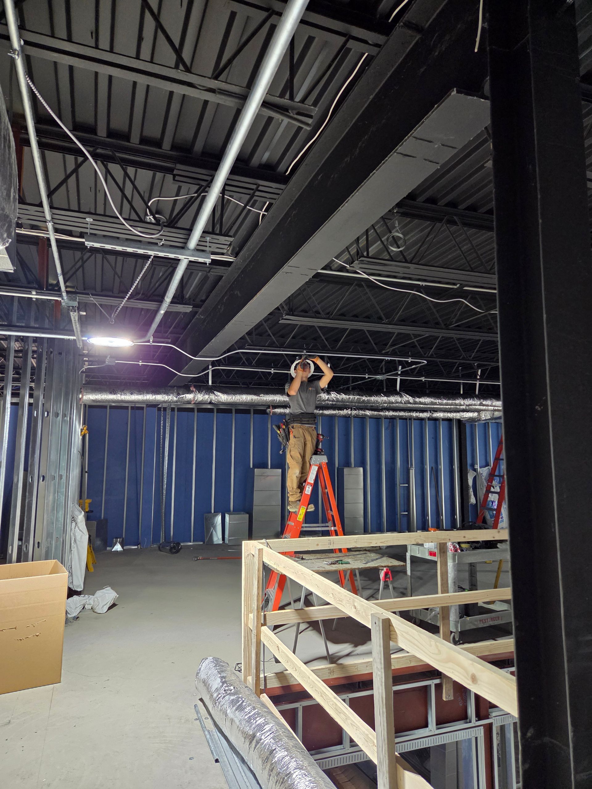 Worker on a ladder installing fixtures in an unfinished room with exposed ceiling and blue wall coverings