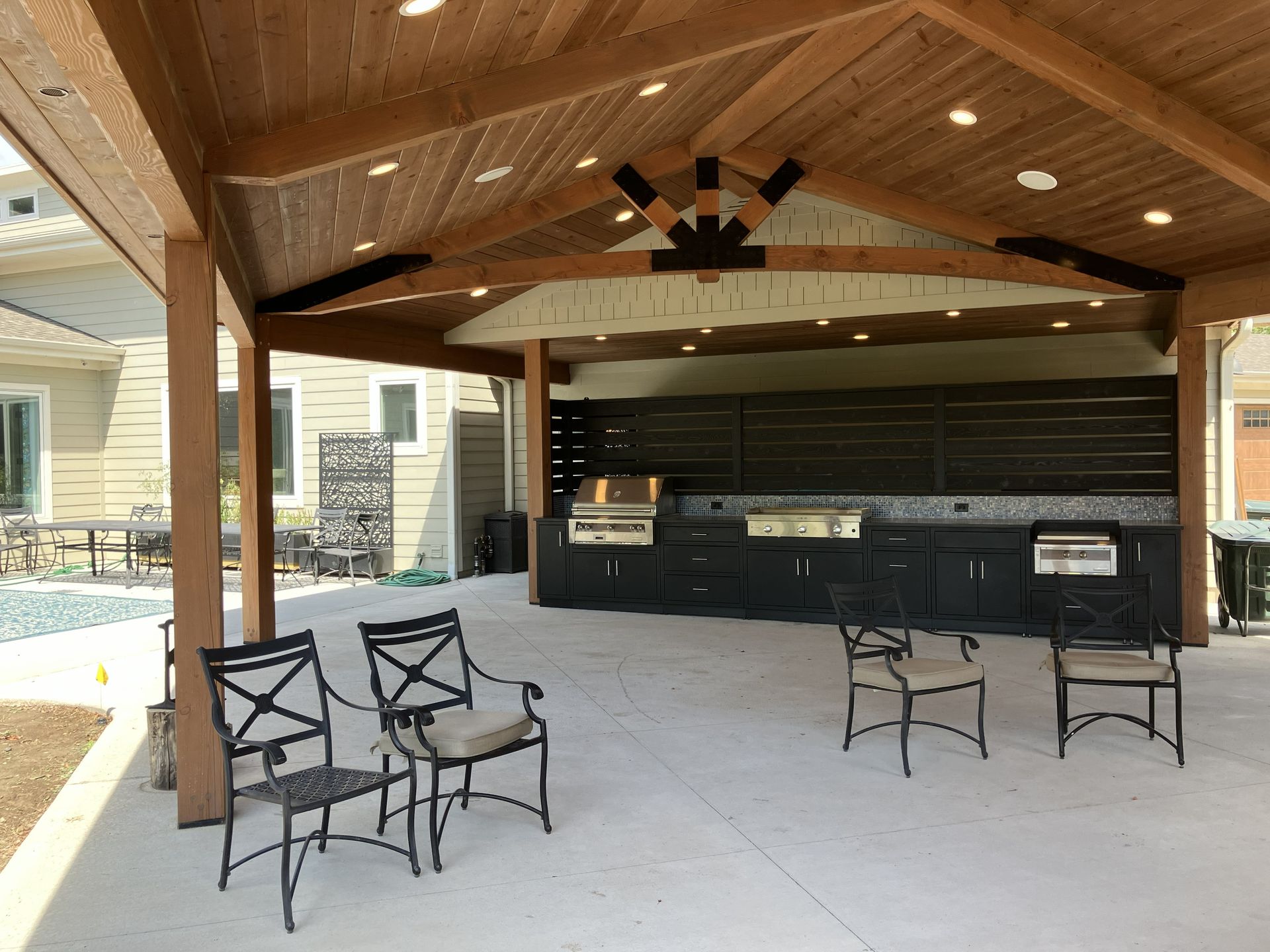 Covered outdoor bar and seating area with wooden ceiling, black stools, and tables on a concrete patio