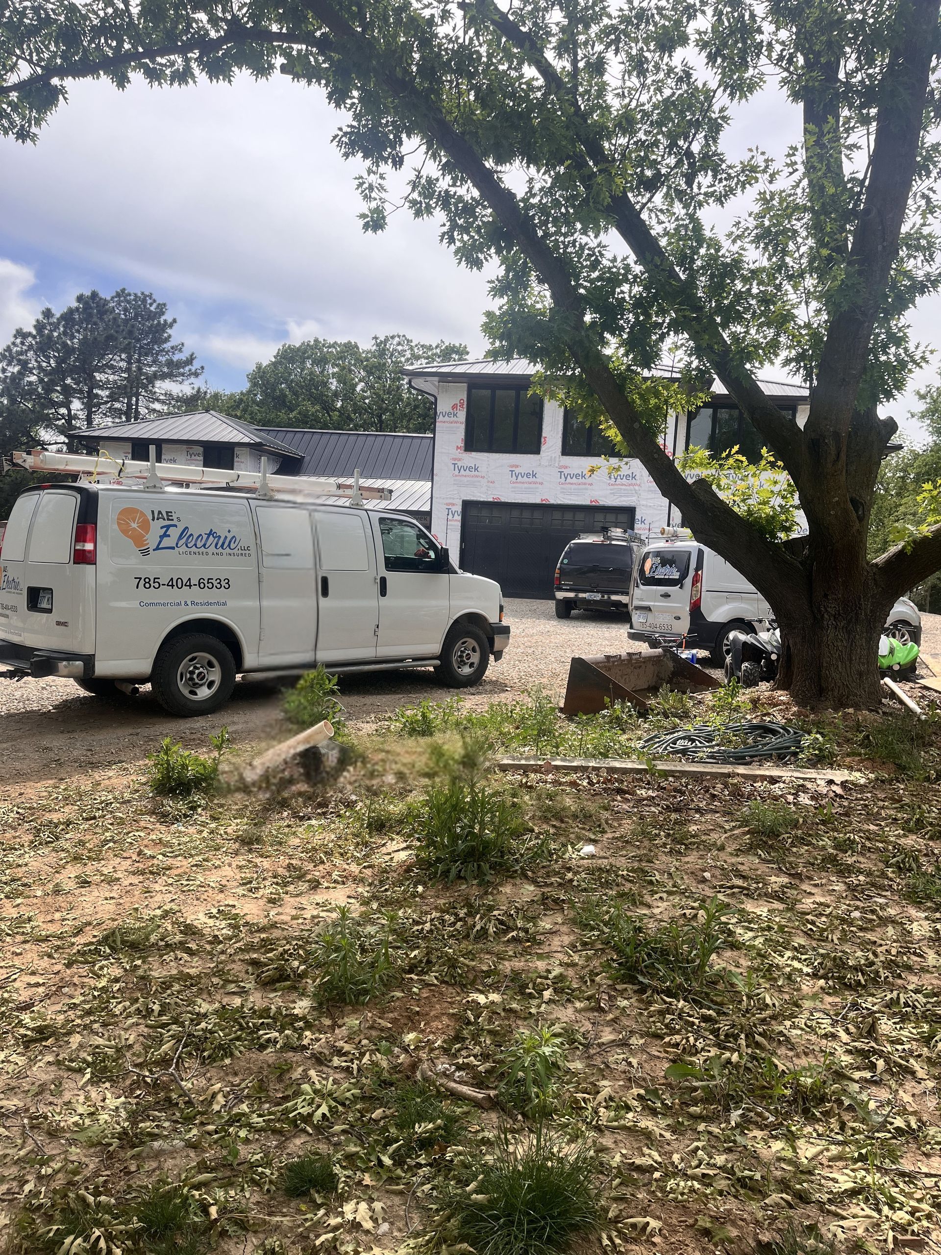 White utility van and other vehicles parked beside a large tree near a house under cloudy sky.