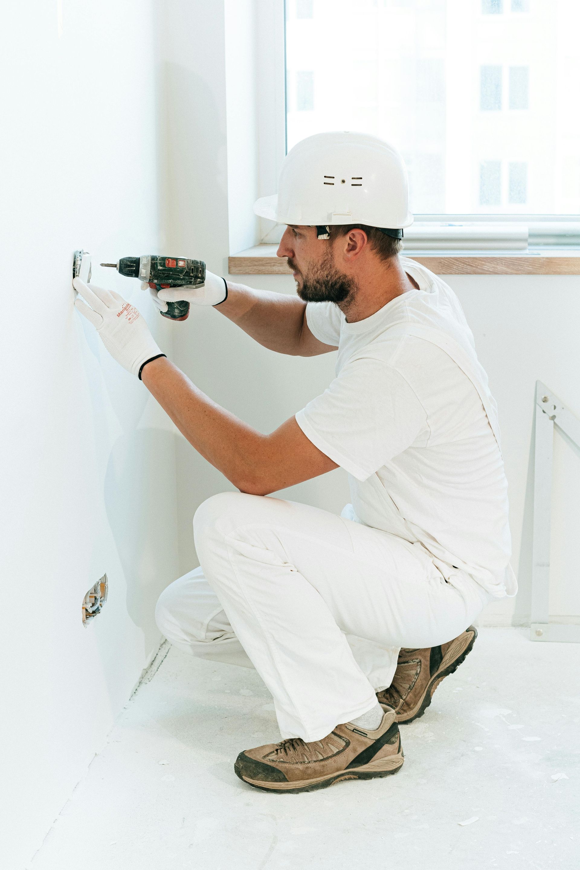 Worker drilling a wall outlet in a bright room, wearing white clothes and a hard hat.