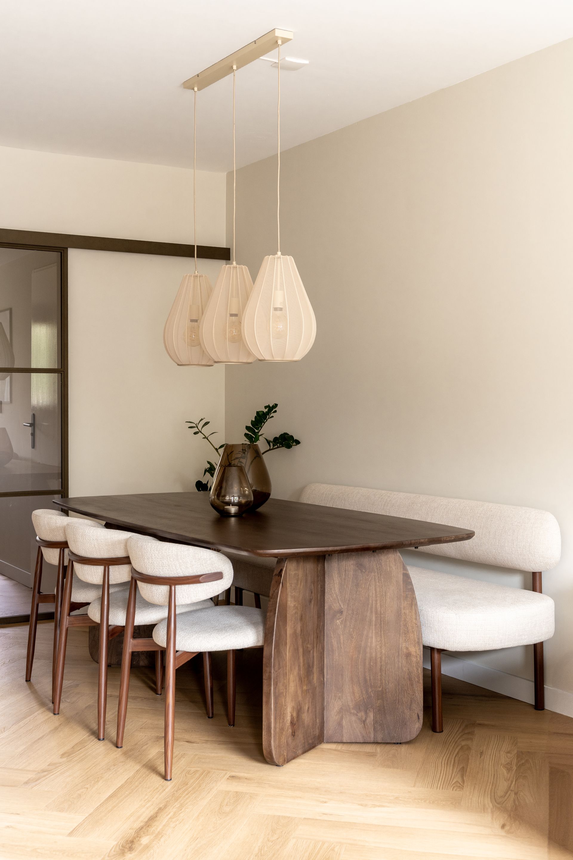 Een eethoek met een donkere tafel, beige hanglampen, beige stoelen en een eetkamerbank op een visgraatparketvloer.