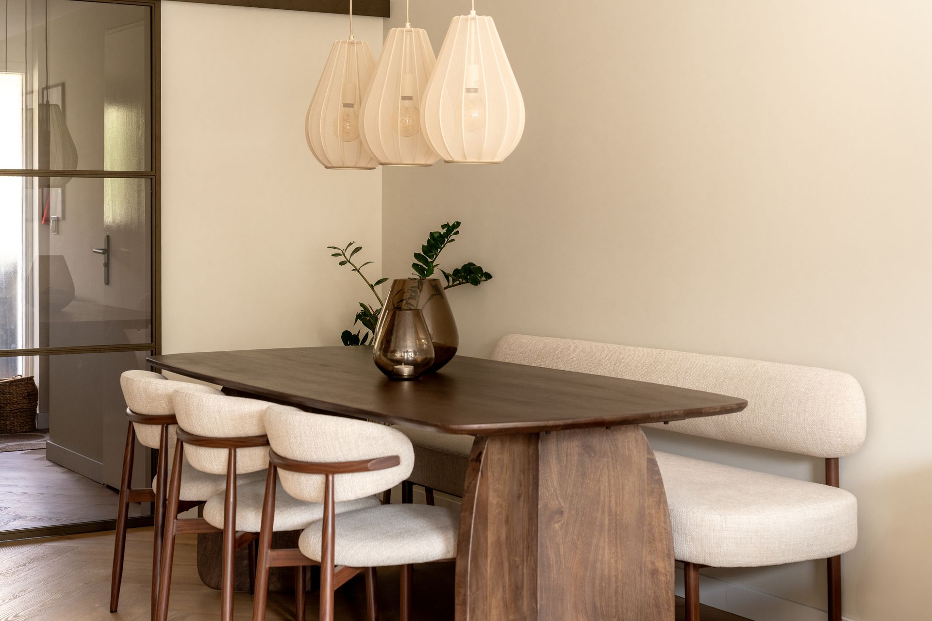 Een eethoek met een donkerhouten tafel, beige stoelen en een eetbank, verlicht door drie hangende pendellampen.