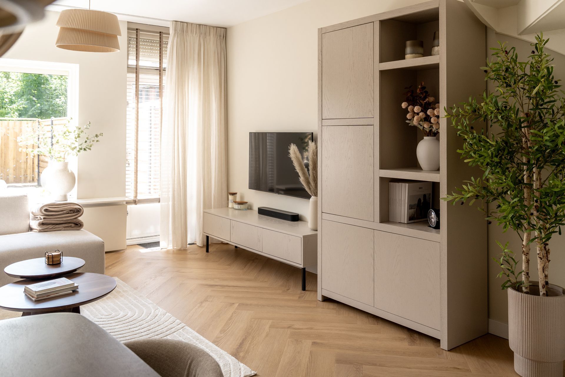 Een lichte woonkamer in neutrale tinten met een lichte visgraatparketvloer, een hoge kast en een aan de muur gemonteerde tv.