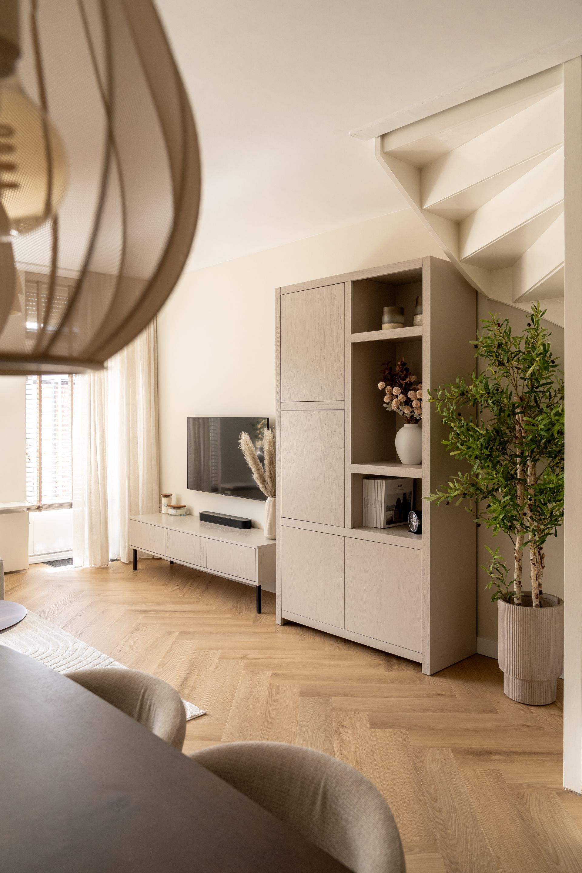 Moderne woonkamer met lichte visgraatvloer, een strakke kast, een aan de muur gemonteerde tv en een potplant bij de trap.
