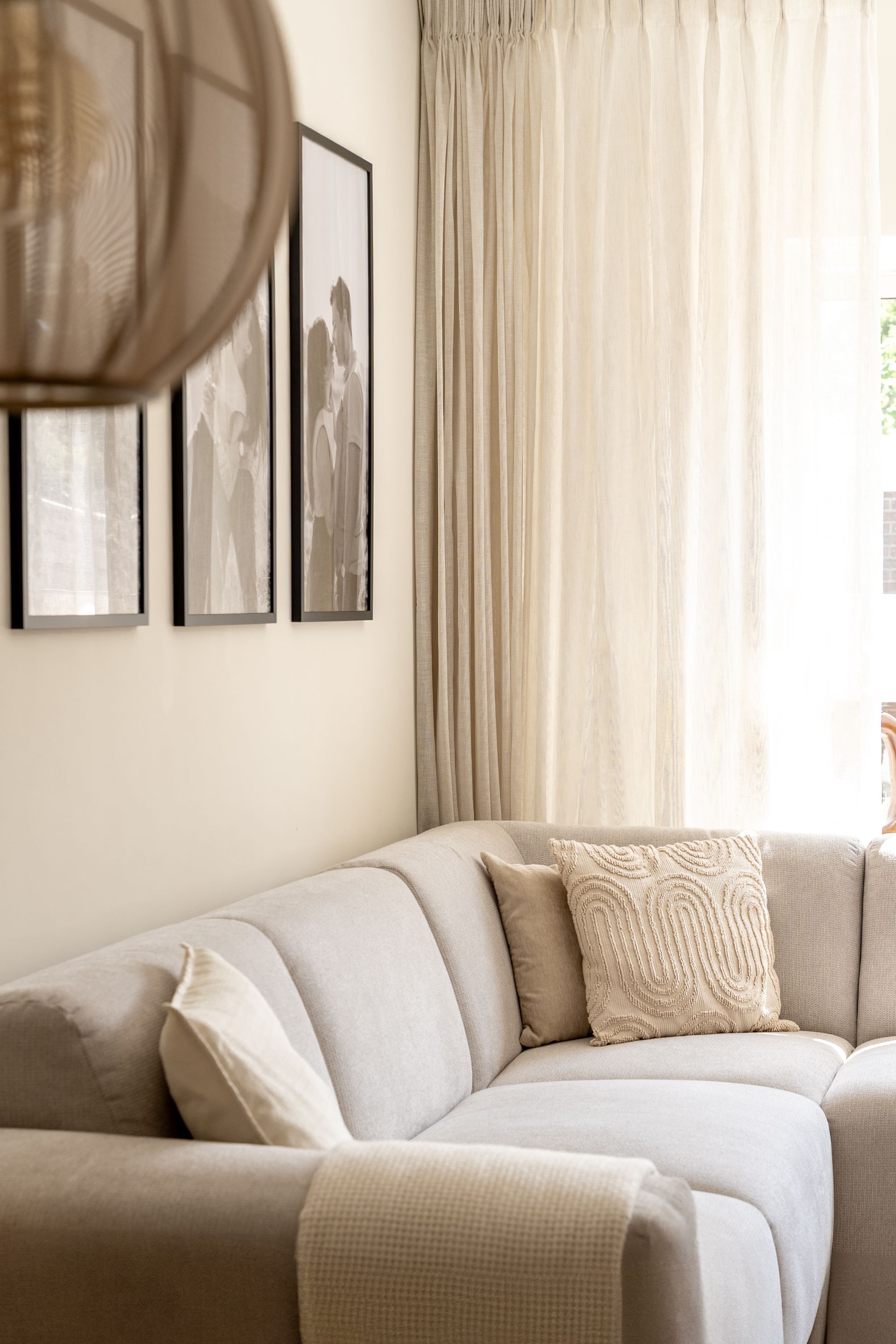 Een beige hoekbank met sierkussens, geplaatst in de buurt van kunst aan de muur en kamerhoge, transparante gordijnen.