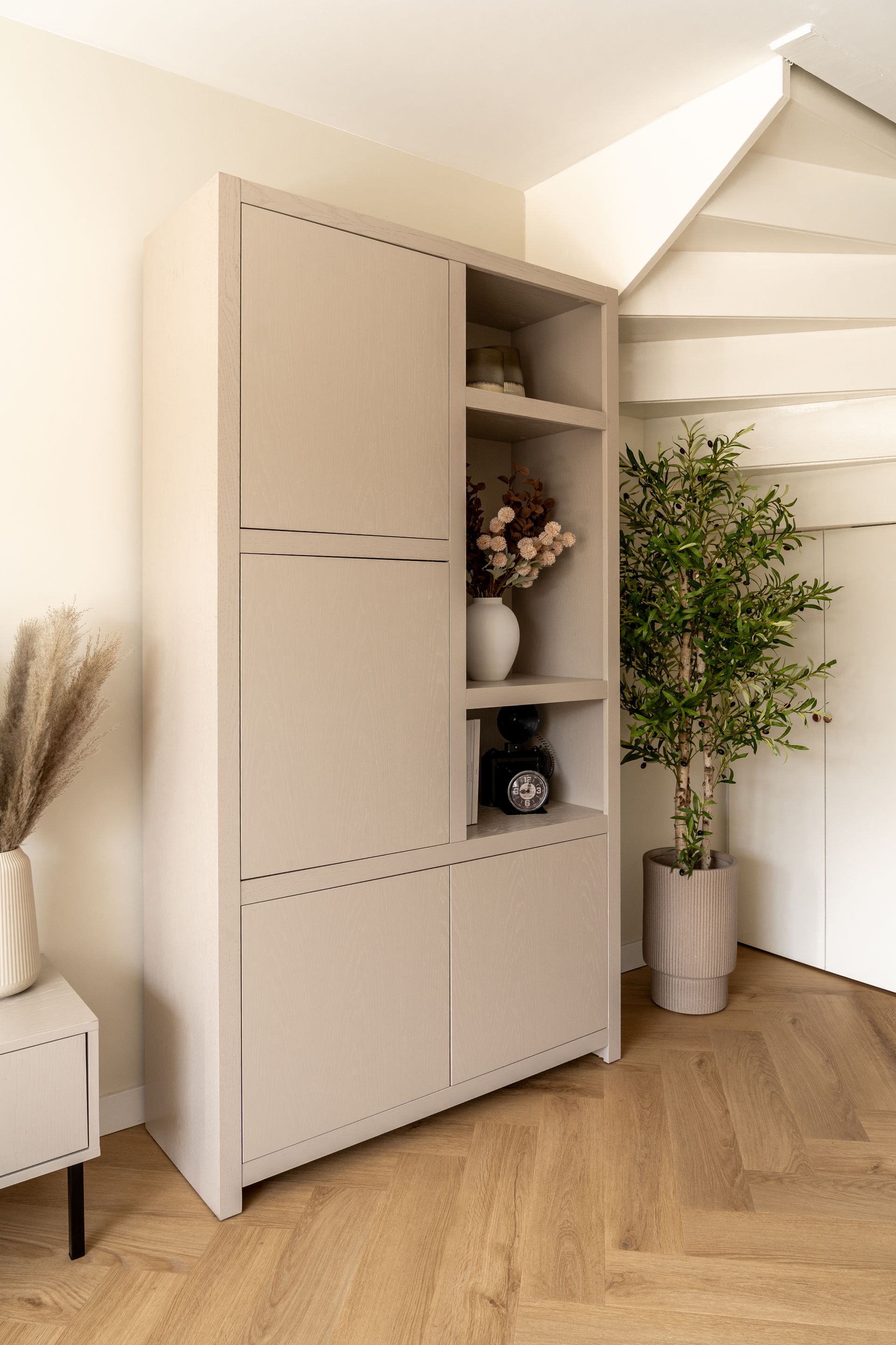 Een beige opbergkast met planken en gesloten deuren staat op een vloer met visgraatmotief naast een groene boom in een pot.