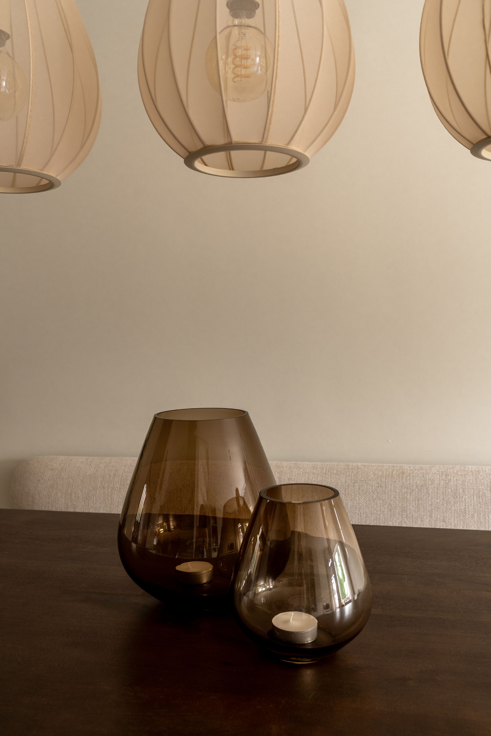 Twee kaarsenstandaards van rookglas staan op een donkere houten tafel onder drie gloeiende, ronde hanglampen.