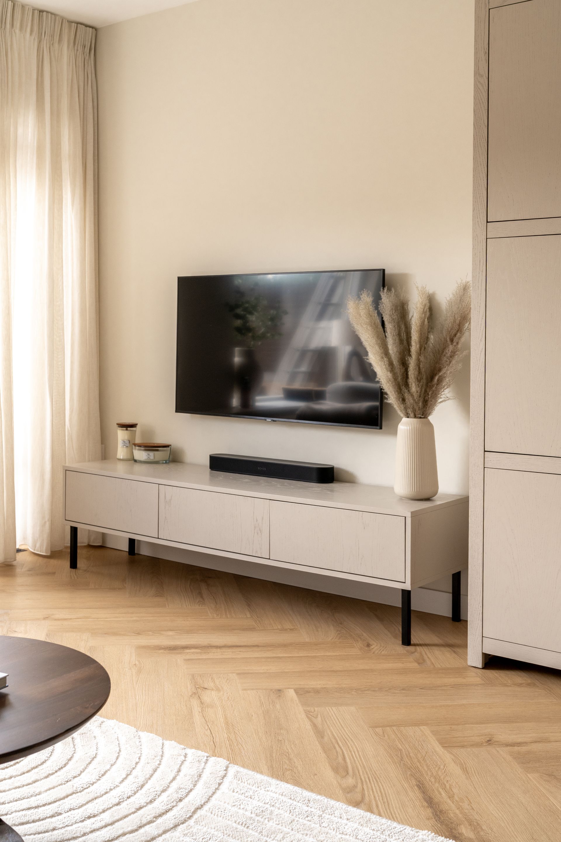 Onder de tv staat een lichtgekleurd tv-meubel, met op de houten vloer een vaas gevuld met gedroogd gras.