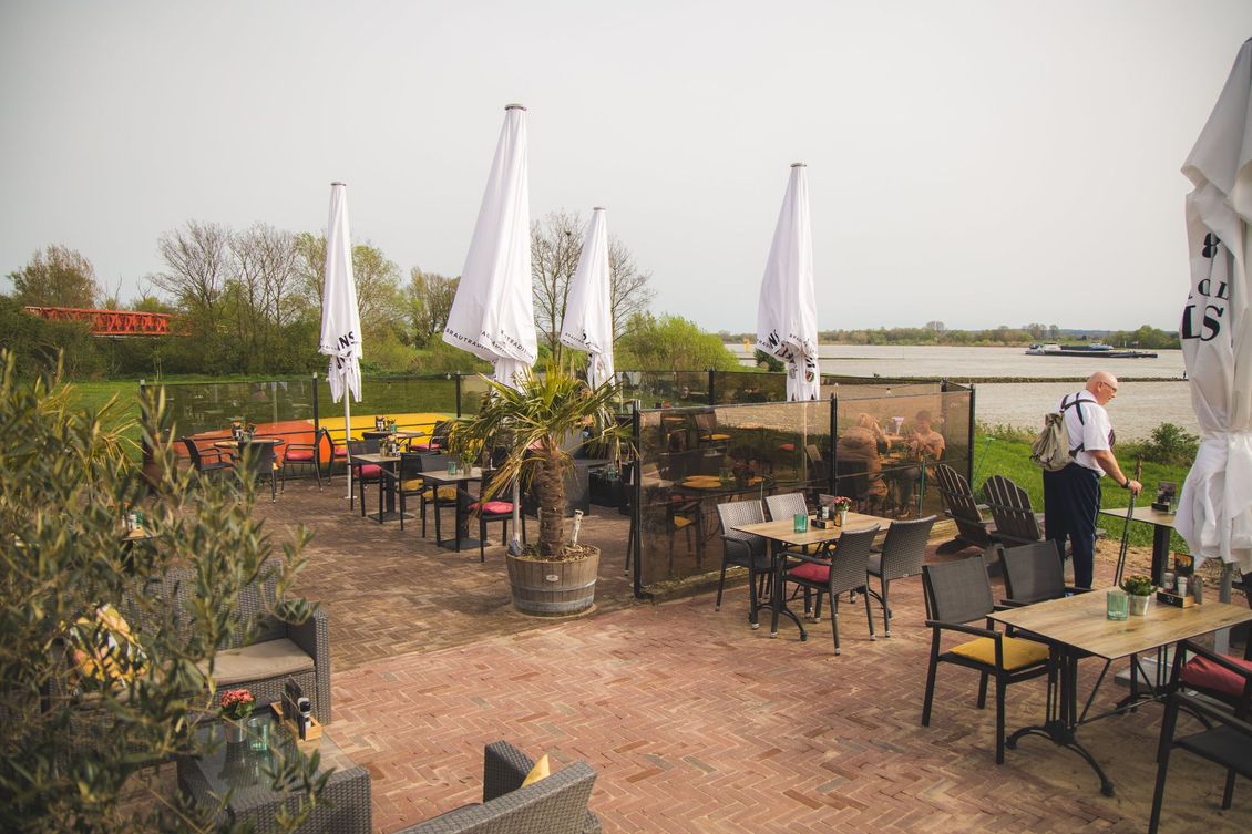 Een terras van een restaurant aan de rivier met diverse tafels, stoelen en gesloten witte parasols op een zonnige dag.