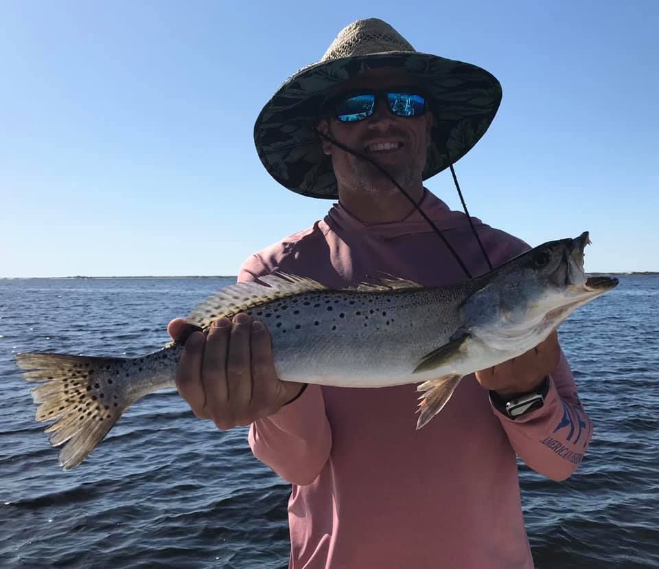 A man wearing a hat and sunglasses is holding a fish in his hands.