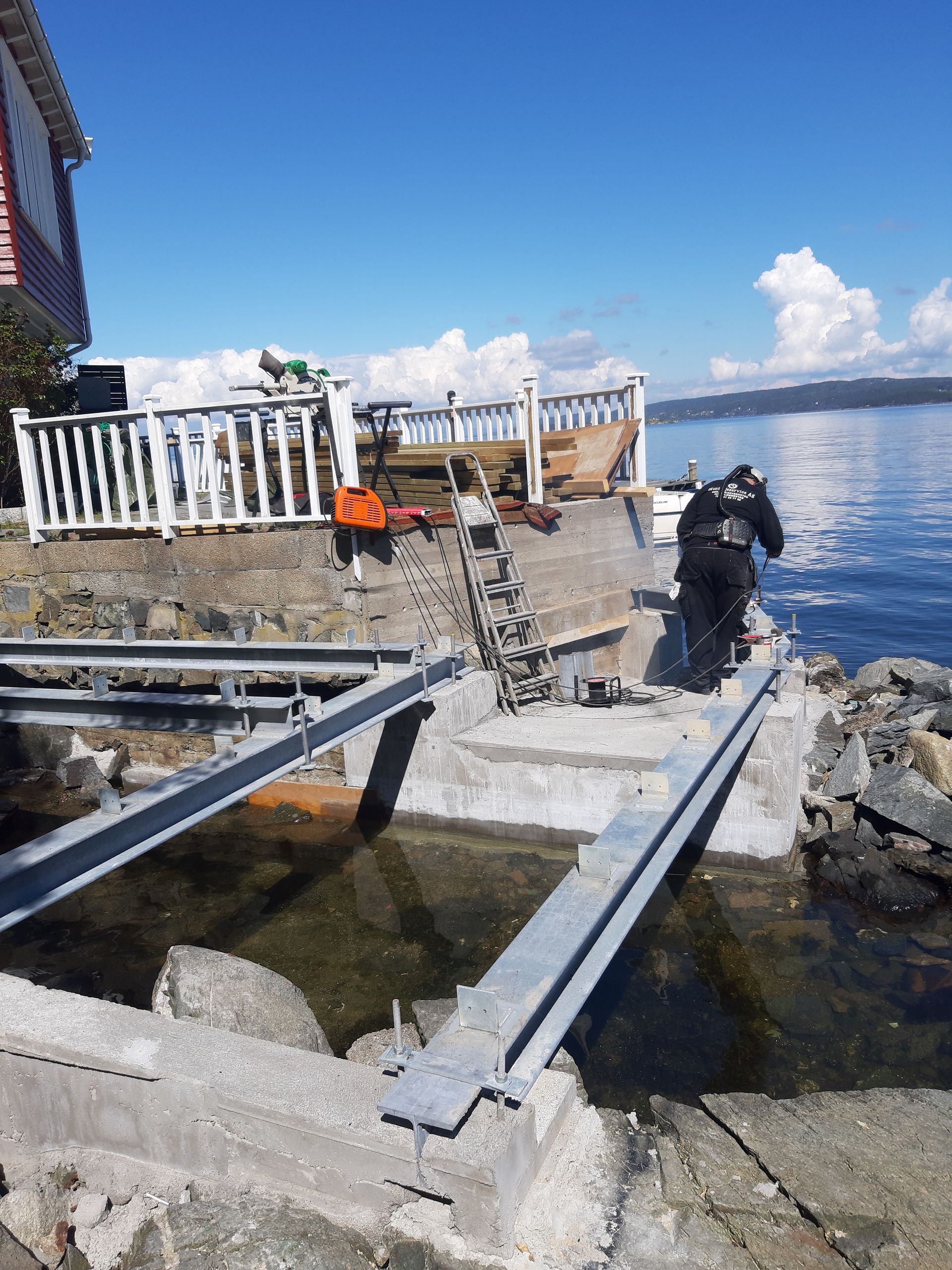 Mann som jobber på en båtrampe ved vannet, nær et hus med terrasse på en solrik dag.